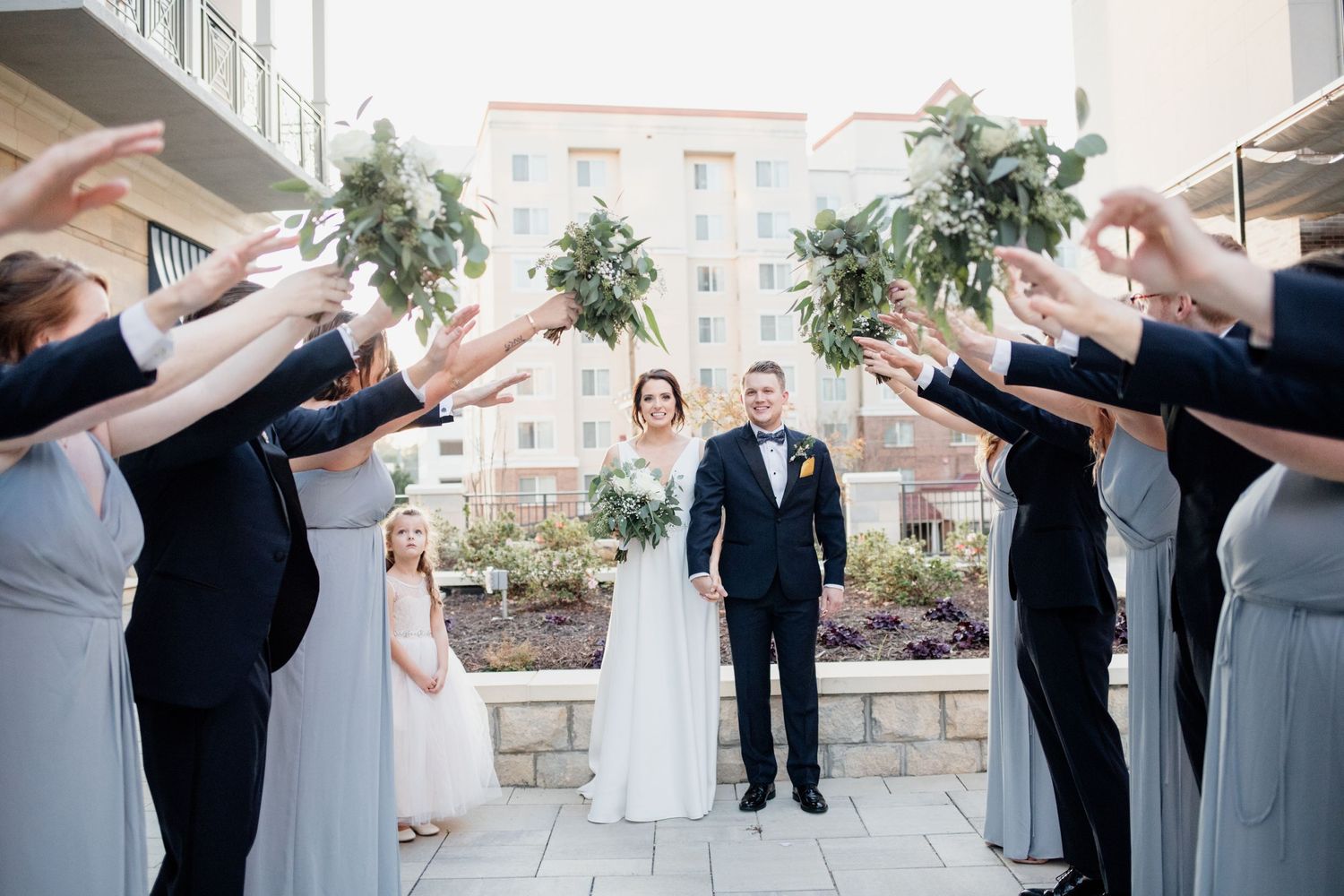 Wedding party forms celebratory archway for couple walking through on stone courtyard.