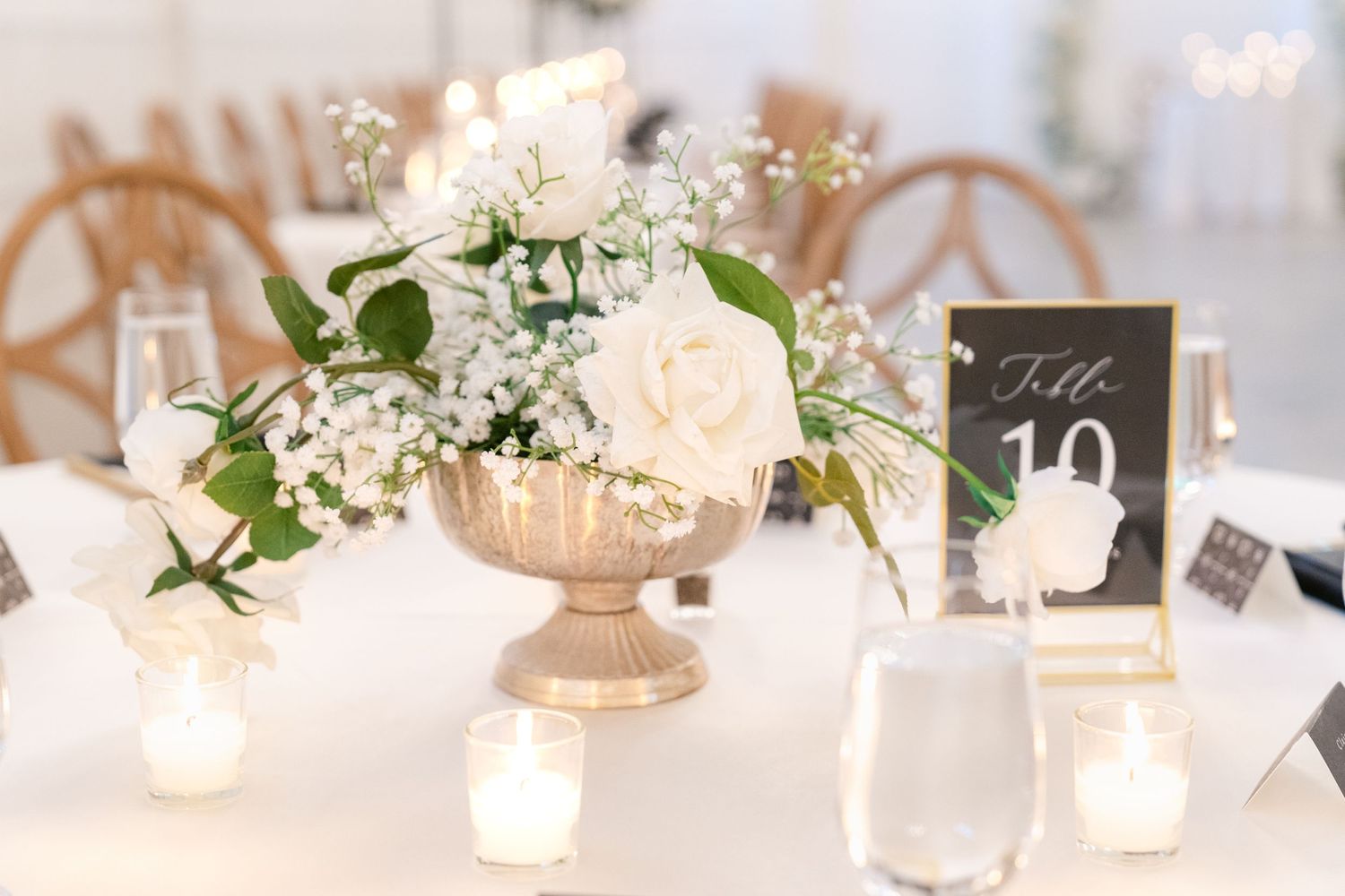 Elegant table setting with white roses and baby's breath in gold pedestal vase surrounded by glowing tea light candles.