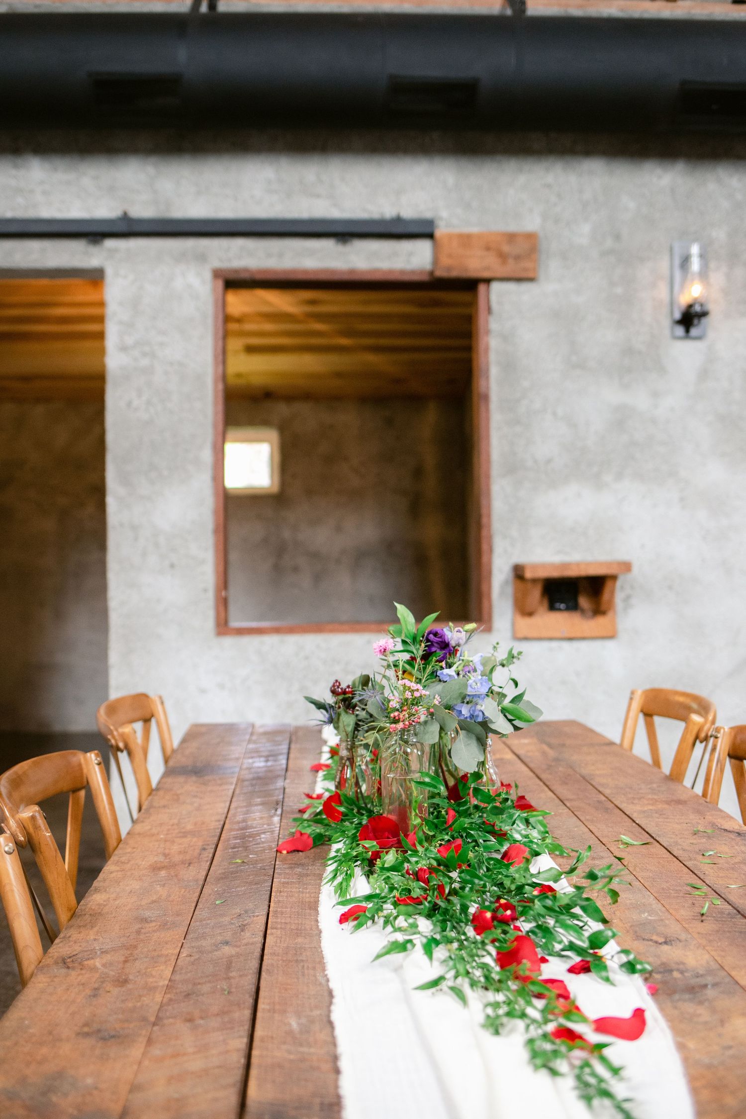 Rustic wooden farm table with floral runner and white linens in industrial space.