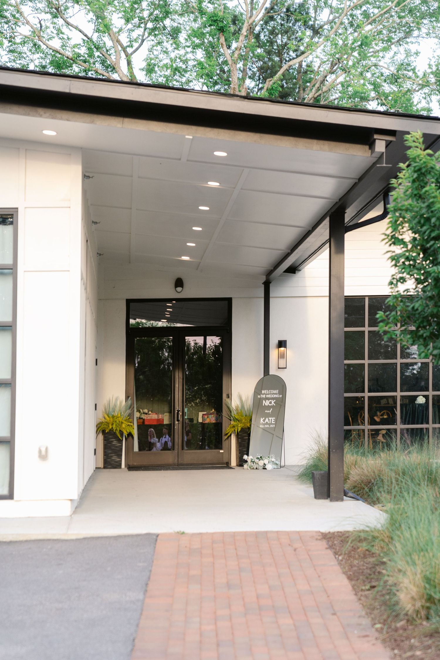 Modern building entrance with covered walkway, white walls, recessed lighting and brick paved path leading to glass doors.