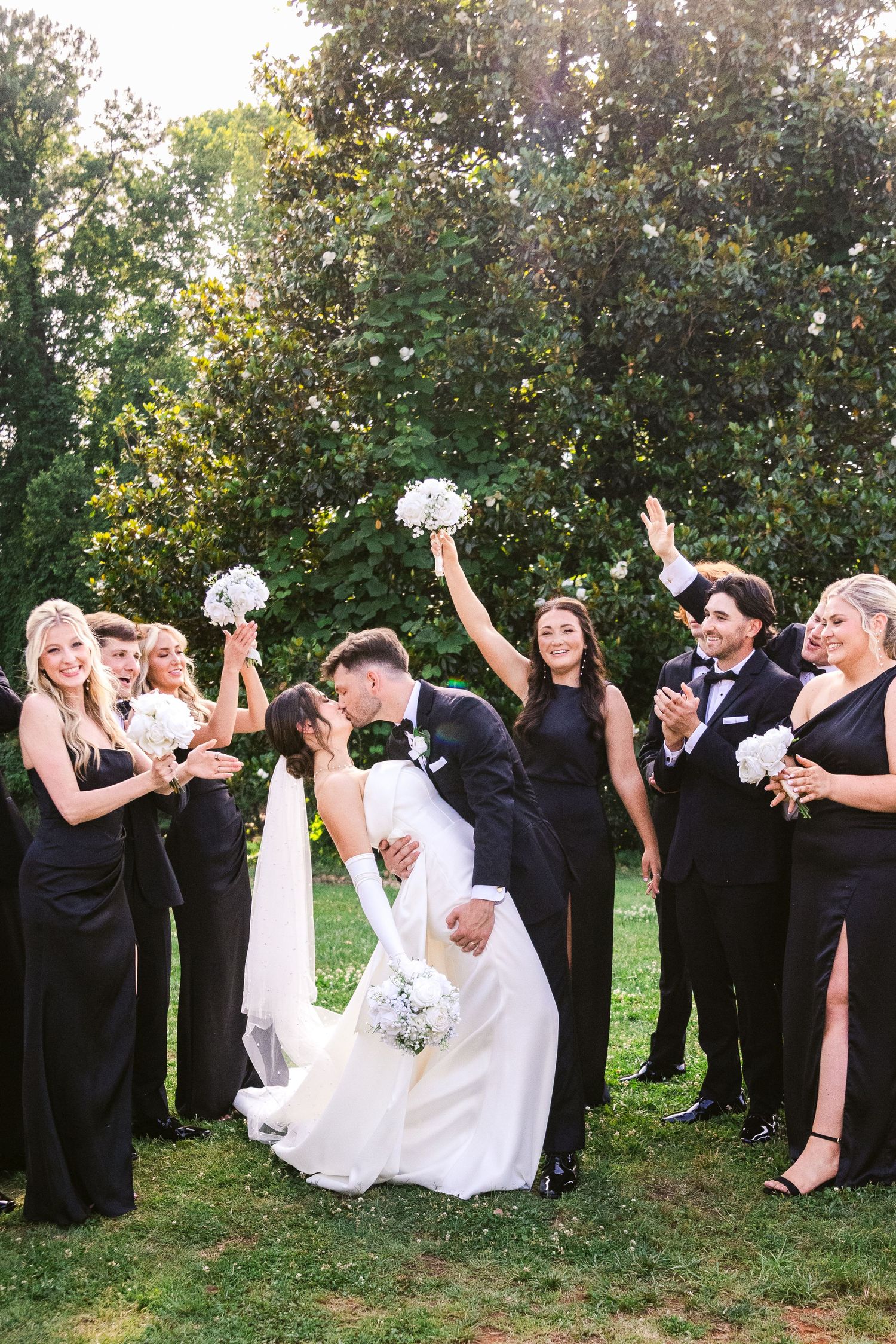 Wedding party in formal attire posing for fun group photos outdoors in a garden setting.