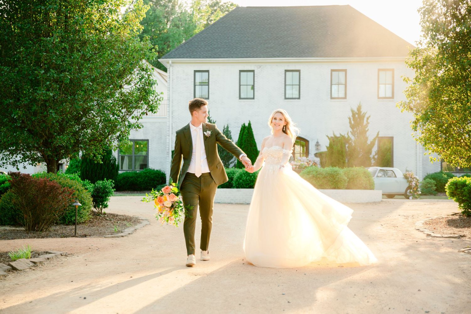 A golden sunset illuminates a wedding couple walking along a path with white buildings and trees in the background.