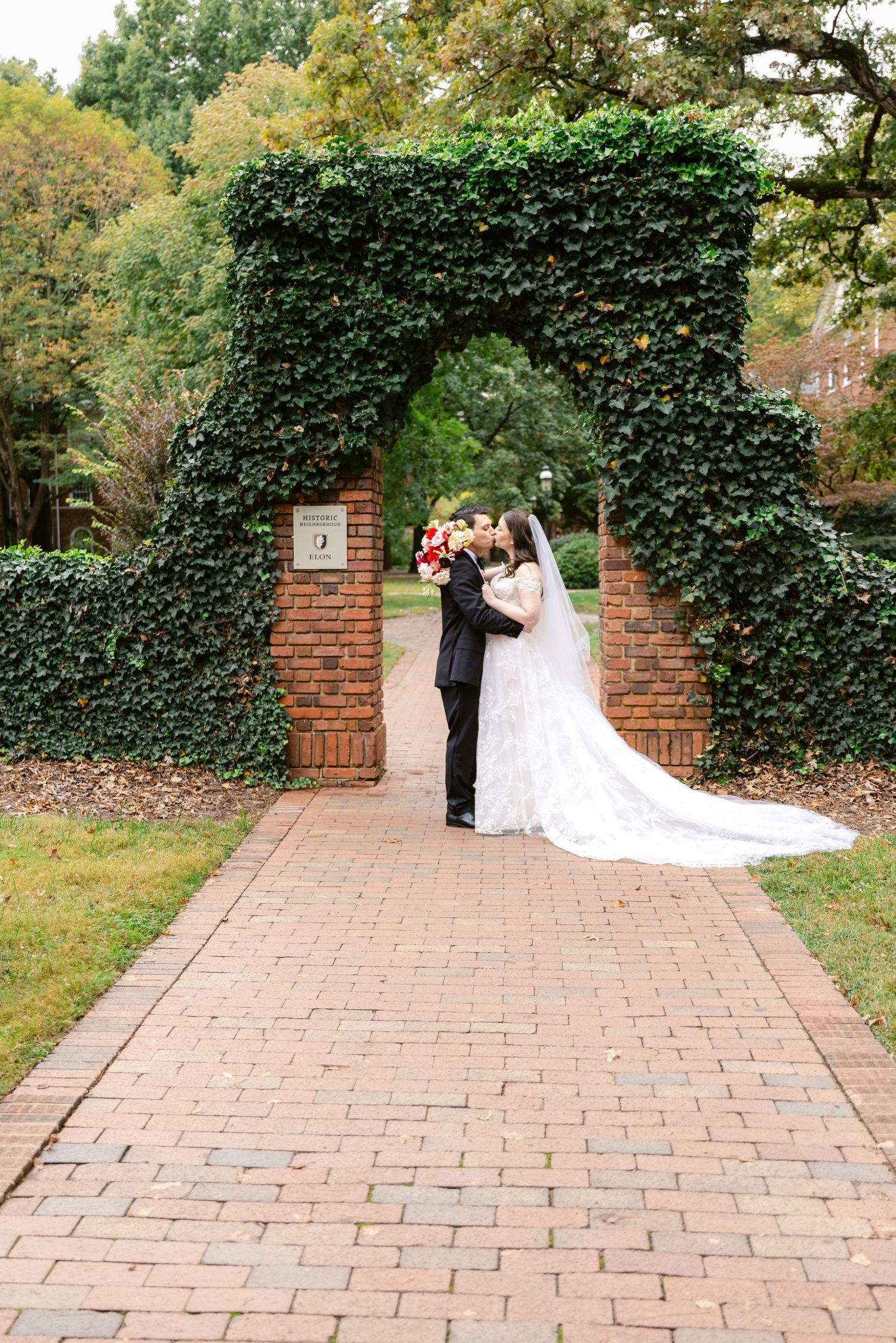 A couple shares a romantic moment under an ivy-covered brick archway on a brick pathway in a garden setting.