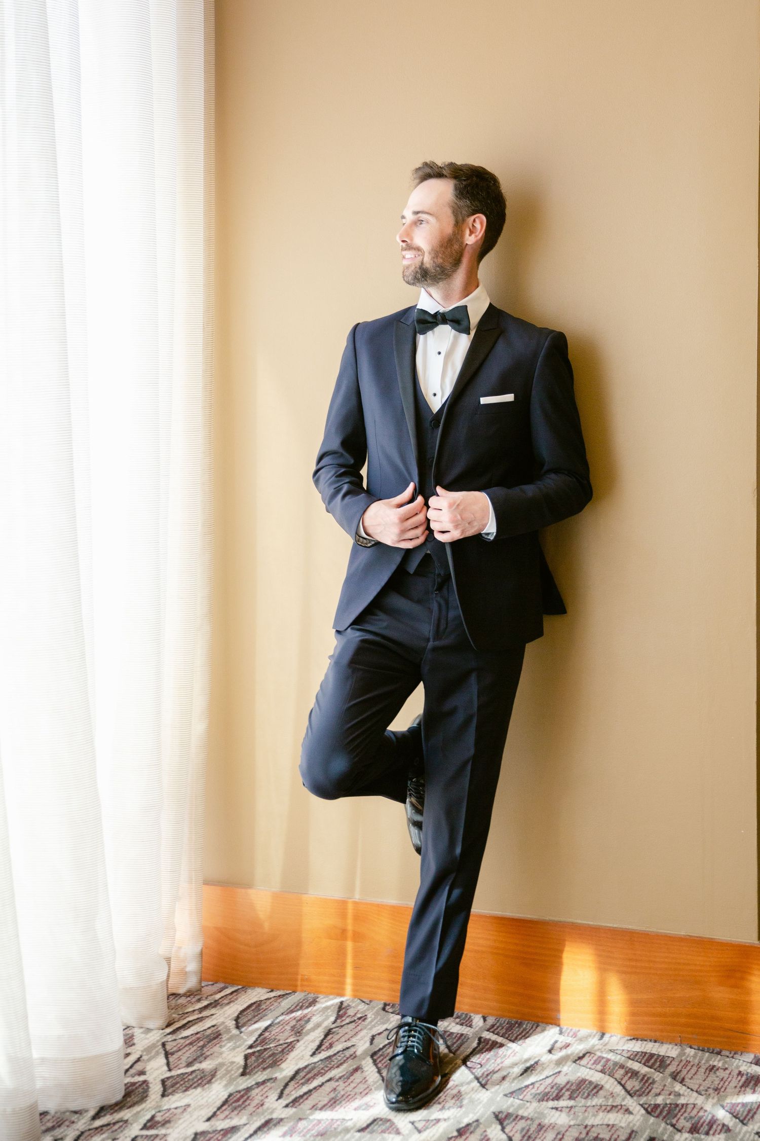 A groom in a classic black tuxedo and bow tie poses elegantly by a window while adjusting his jacket.