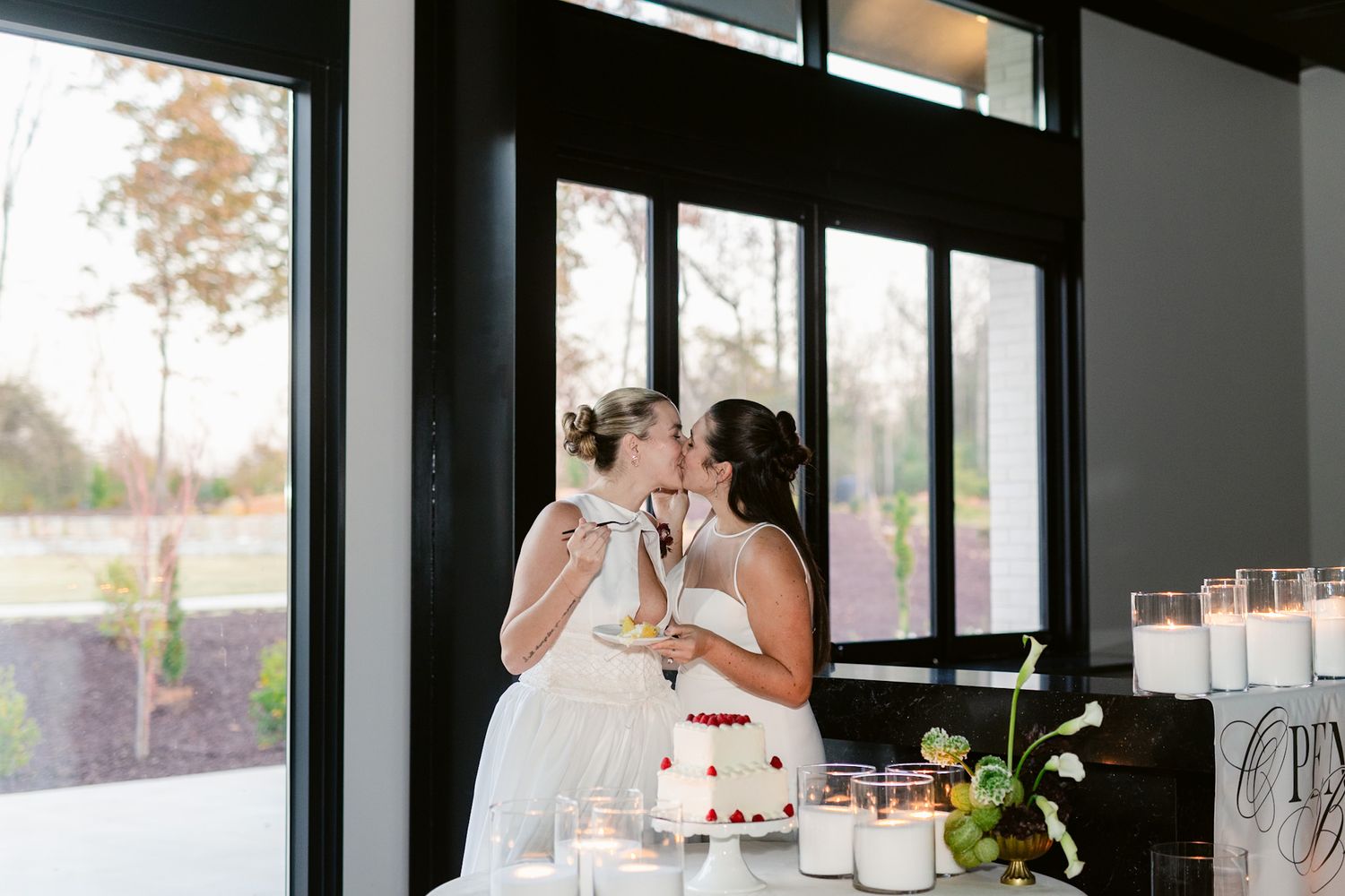 Wedding couple shares a romantic kiss during their reception in an elegant venue with large windows and natural light.
