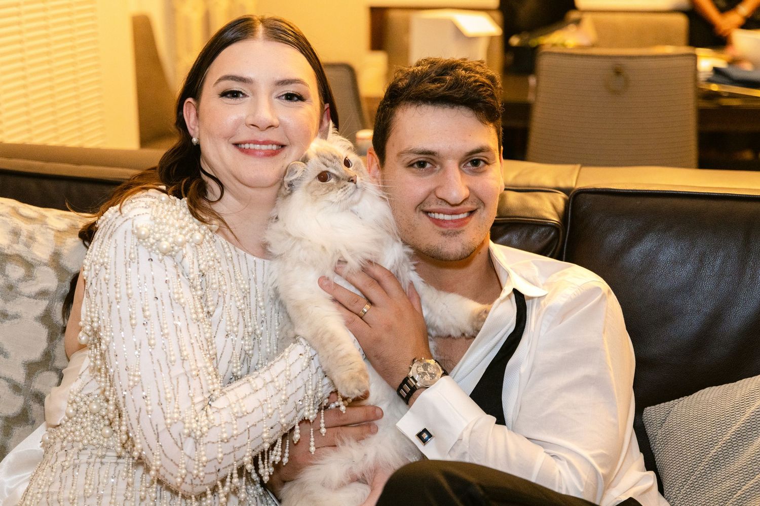 A couple sits on a couch holding a fluffy cream-colored cat while wearing cozy white sweaters.