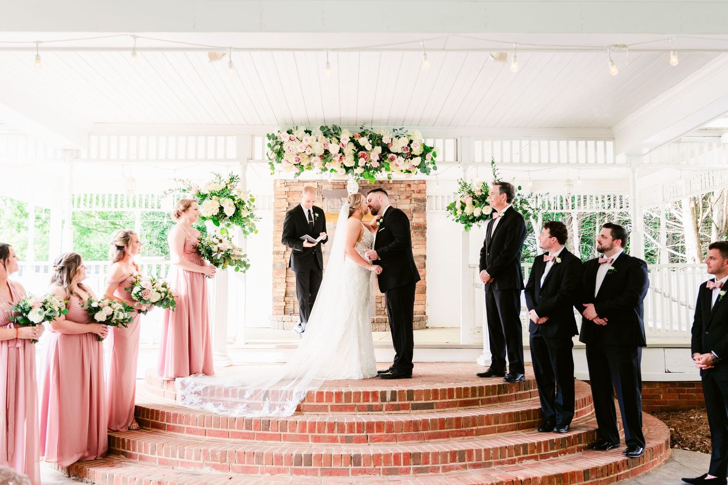 Elegant outdoor wedding ceremony with pink bridesmaids and black-suited groomsmen on brick steps.