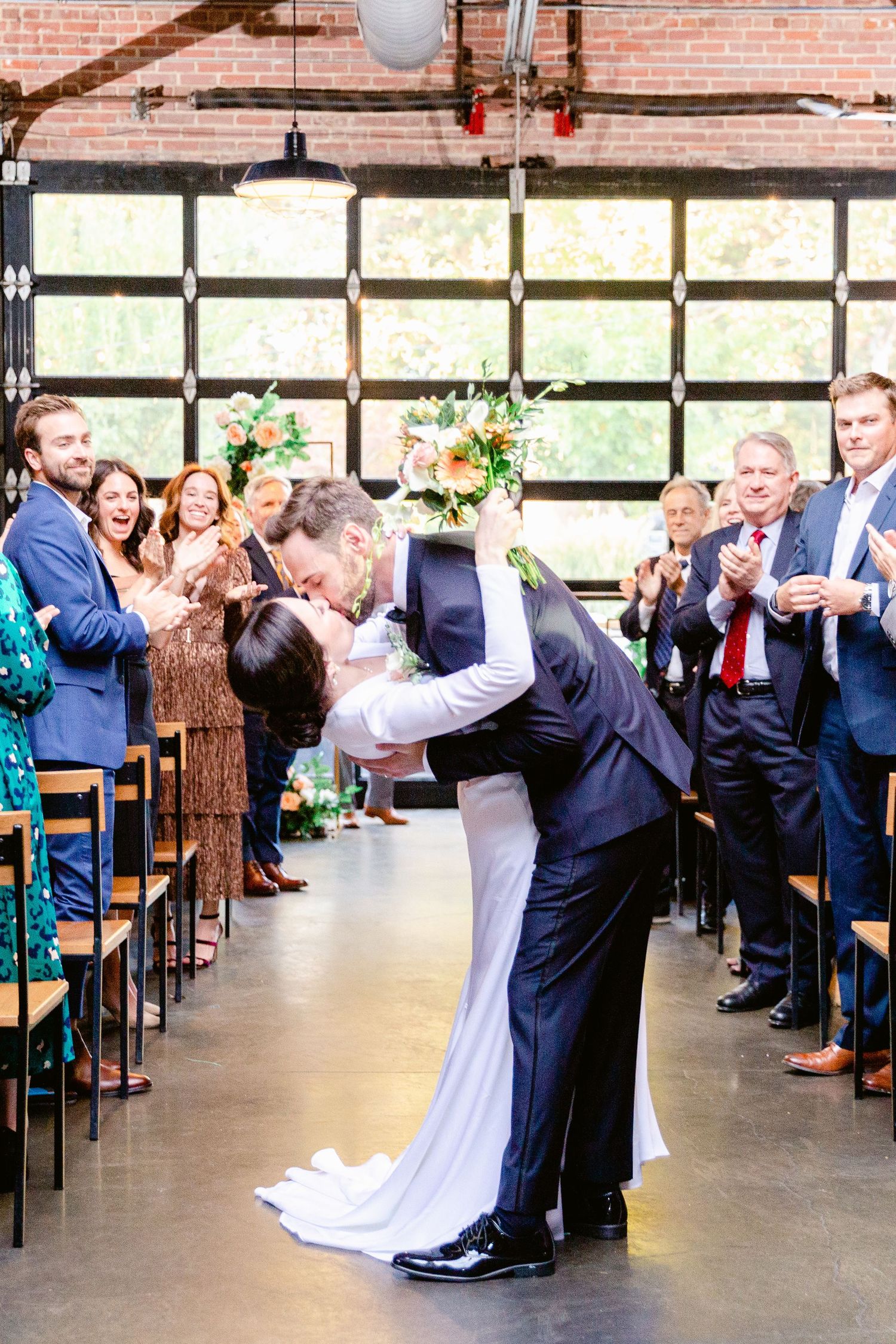 Newlyweds execute an elegant dance dip move in a rustic brick-walled reception venue with large windows.