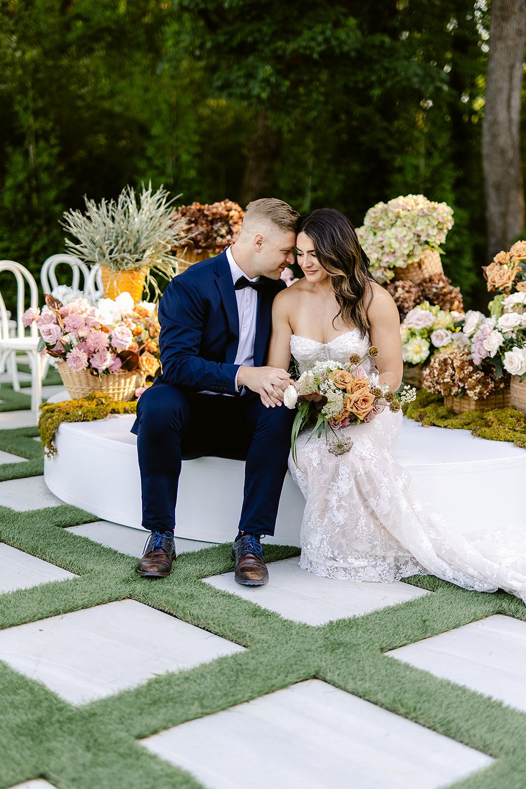 Wedding couple sitting on white platforms arranged on grass with floral decorations and outdoor reception setup.