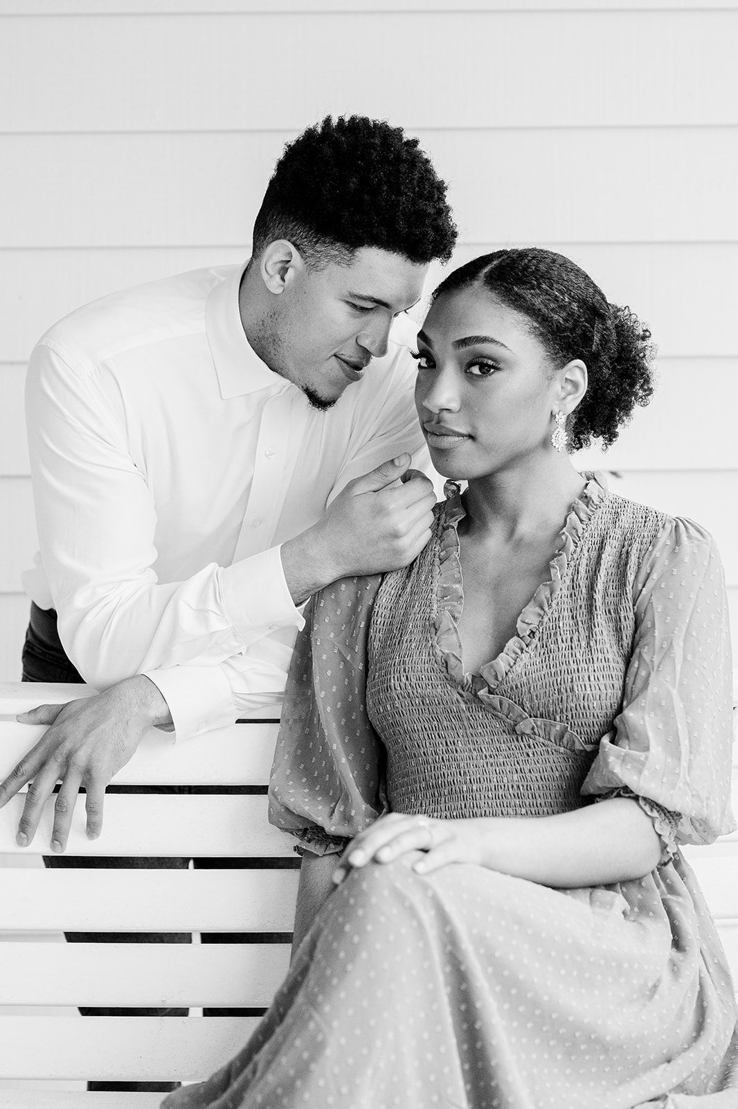 A romantic black and white portrait of a couple in elegant attire sitting close together on a white bench.