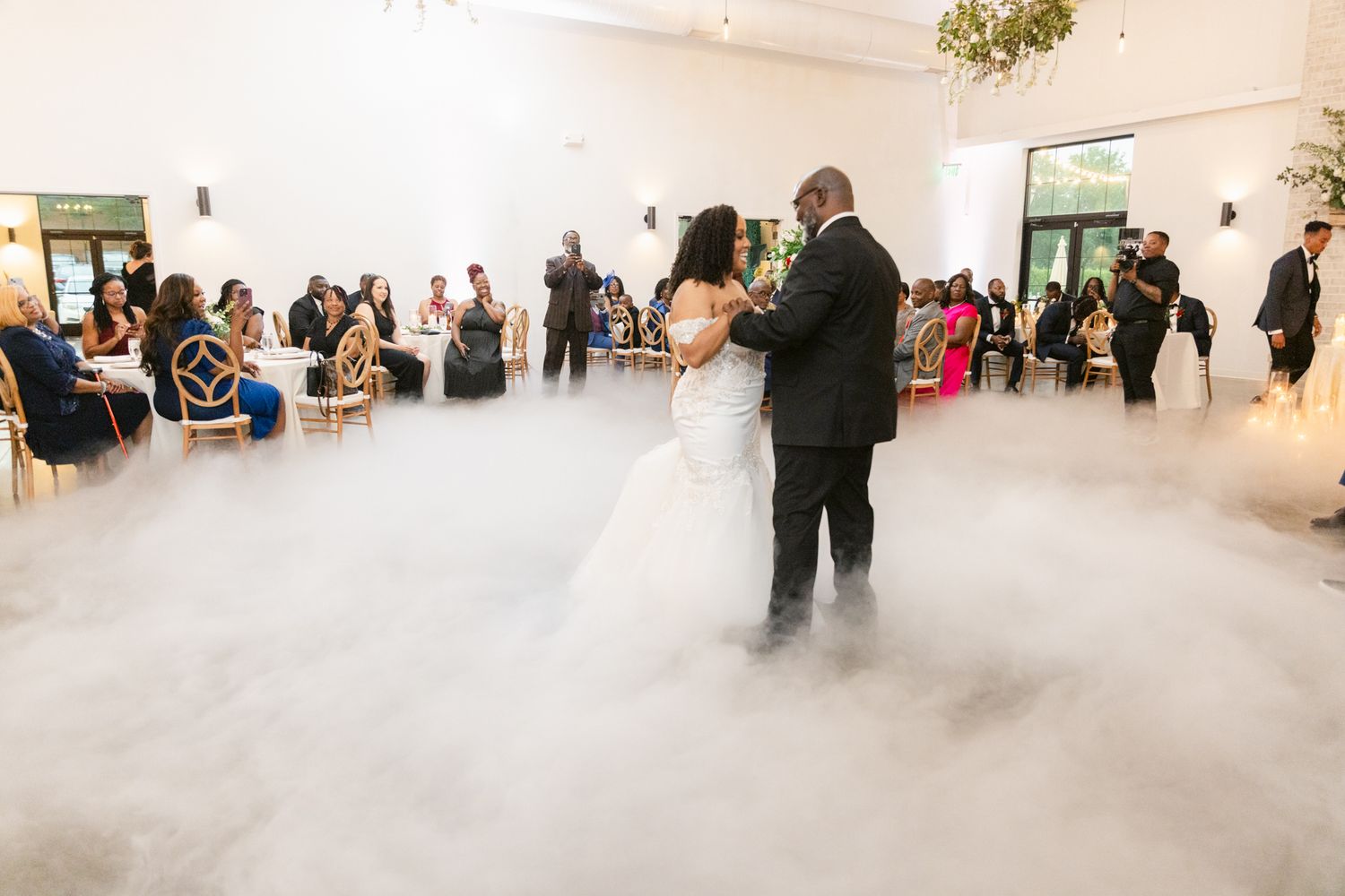 Wedding guests dancing and celebrating in an elegant white reception venue with high ceilings.