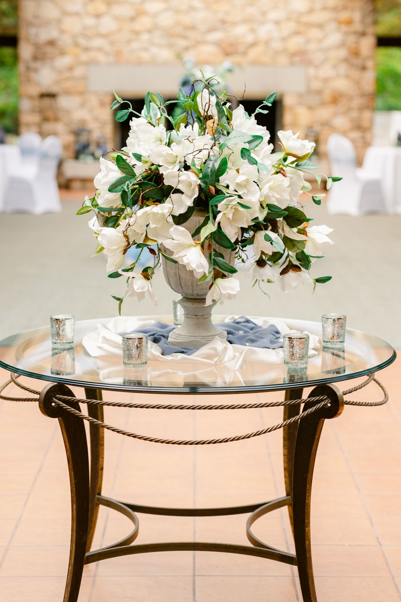 Elegant glass table with white floral centerpiece and metallic rope details displayed at an outdoor event setting.