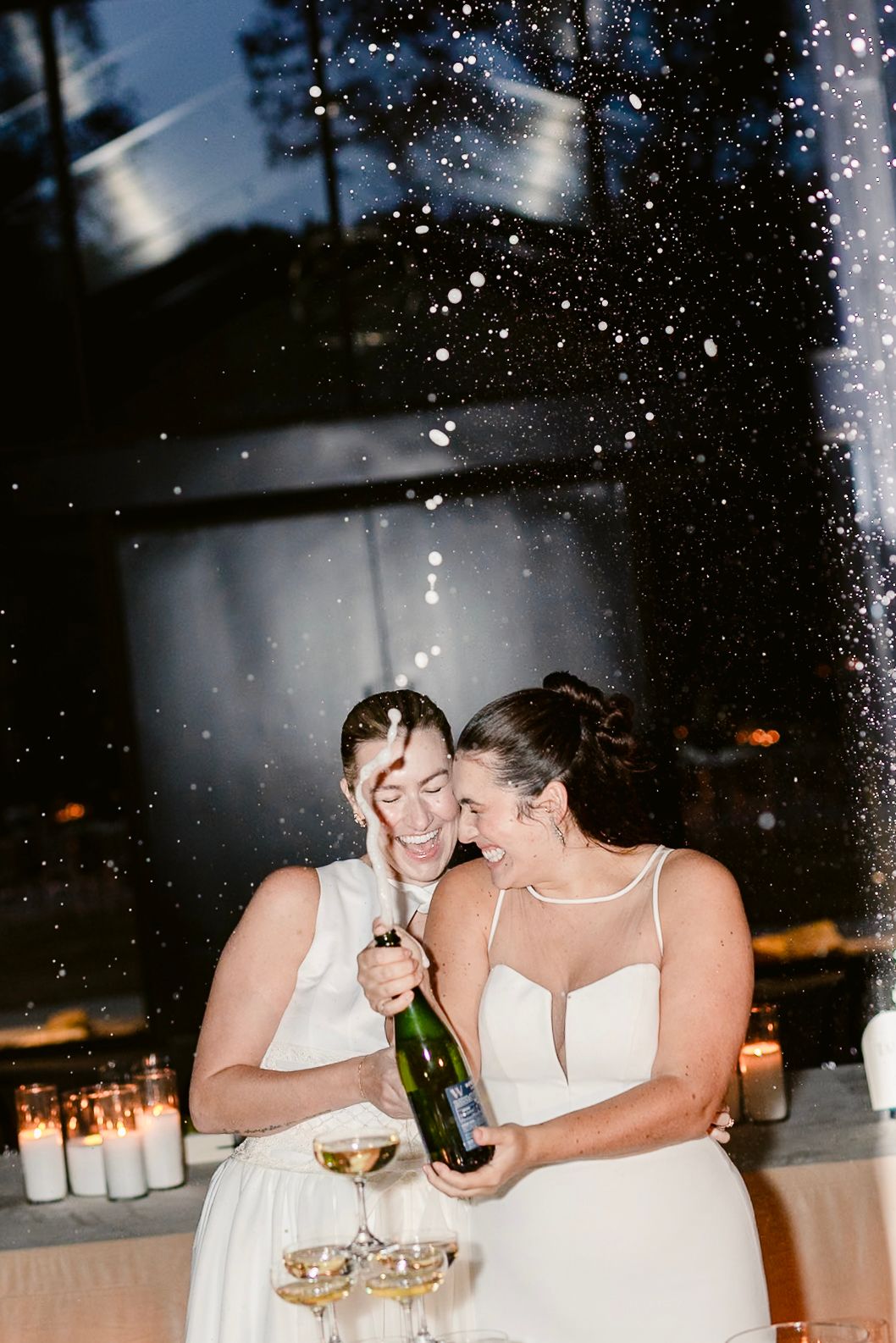 Two people in white dresses share a joyful moment during a wedding celebration with champagne and falling confetti.
