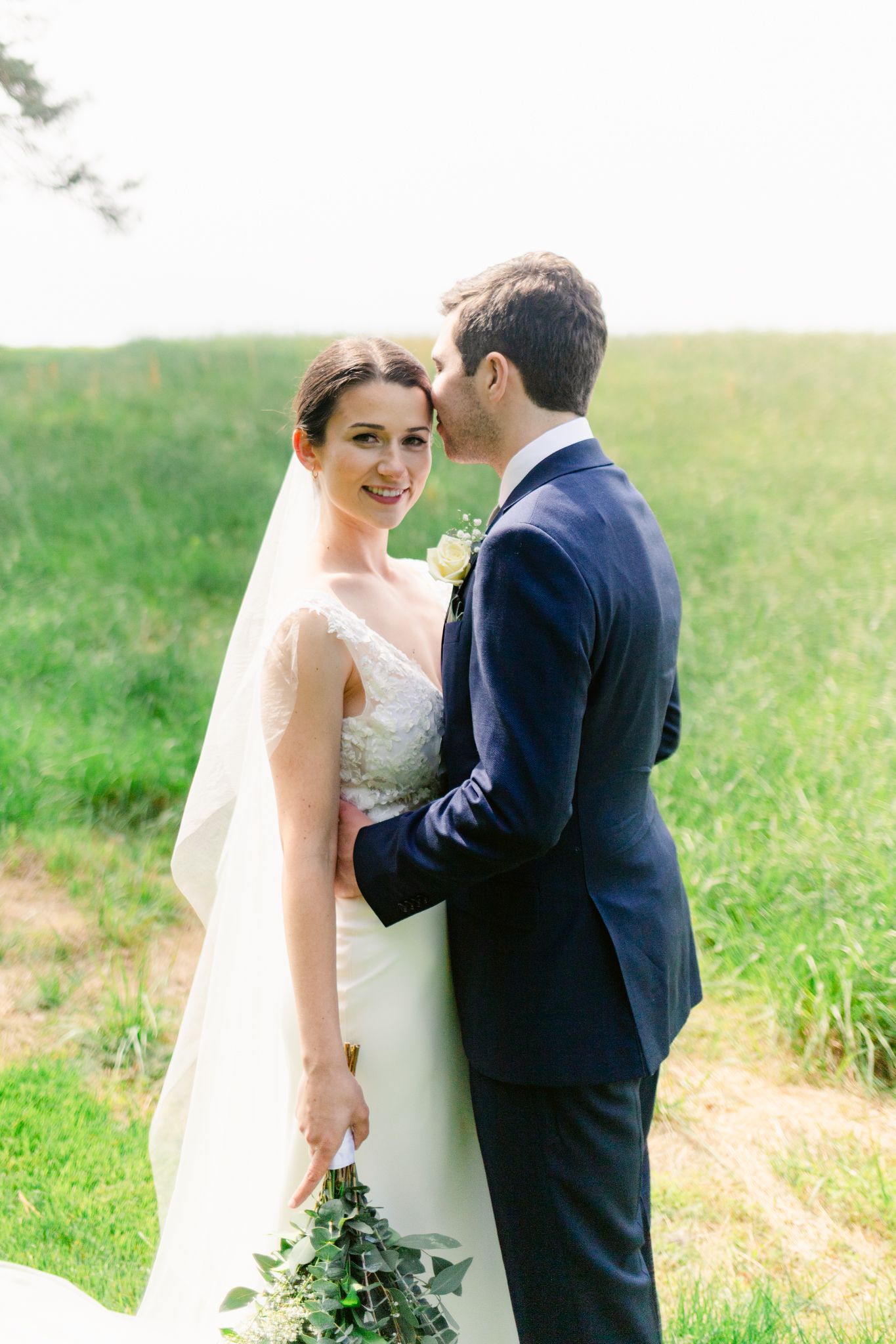 A bride and groom share an intimate moment in a sunlit grassy field on their wedding day.