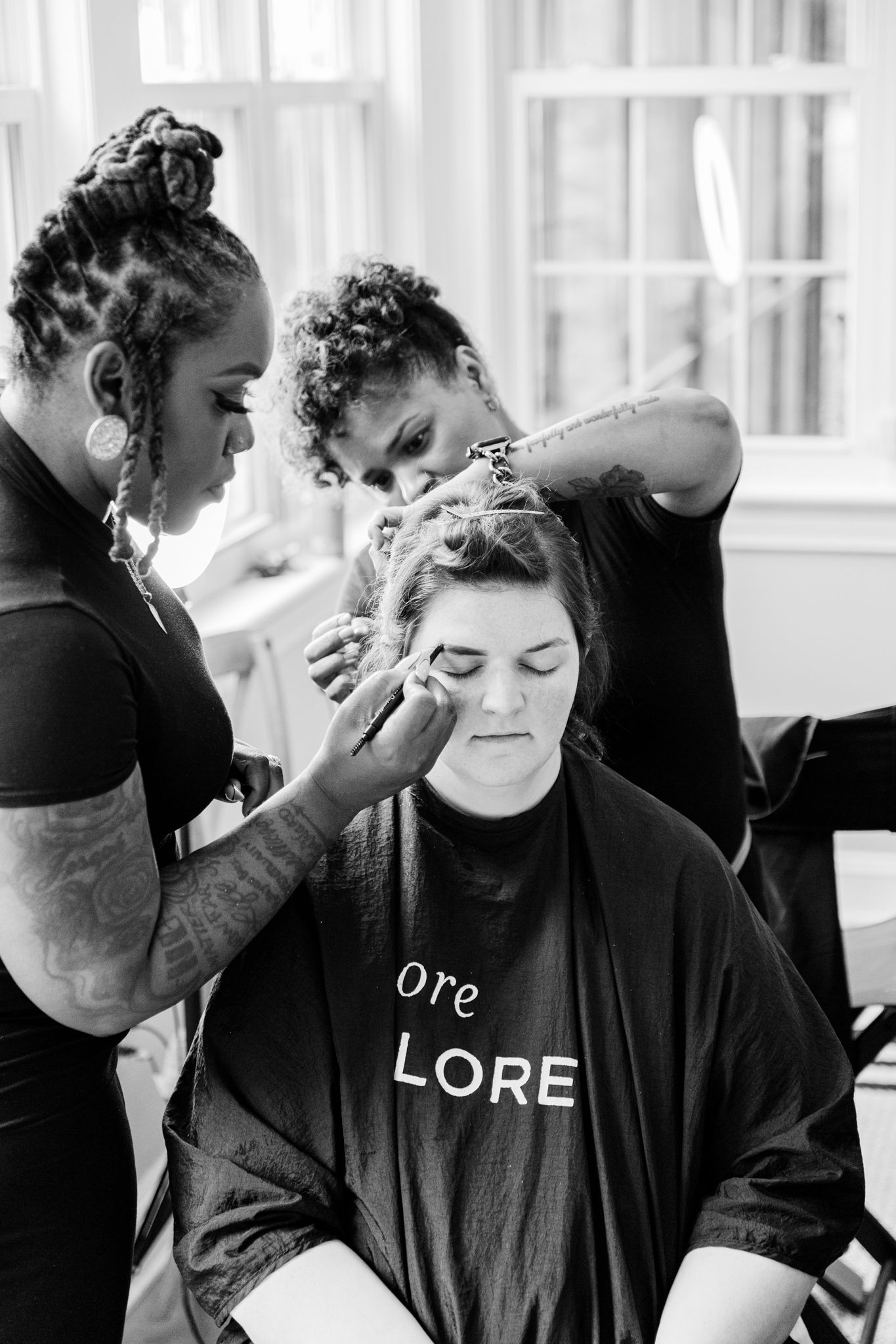 Black and white photo of makeup artists working on a client wearing a LORE shirt in a bright studio setting.