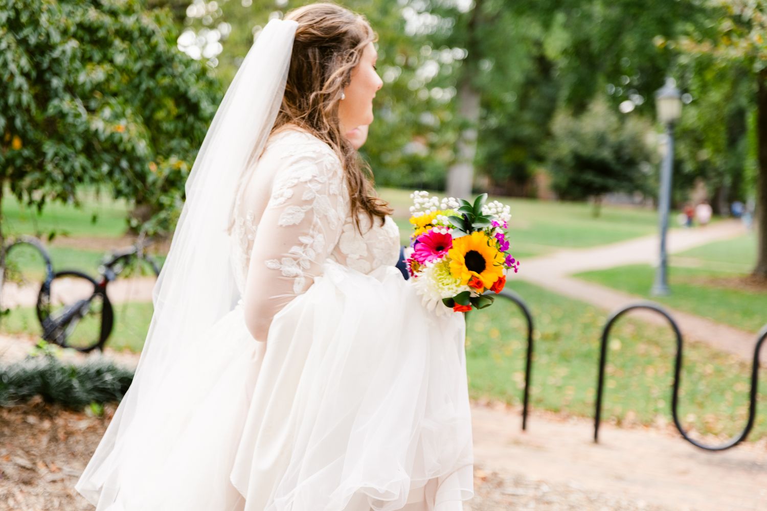 A person in a flowing white dress walks through a park pathway holding a vibrant sunflower bouquet.