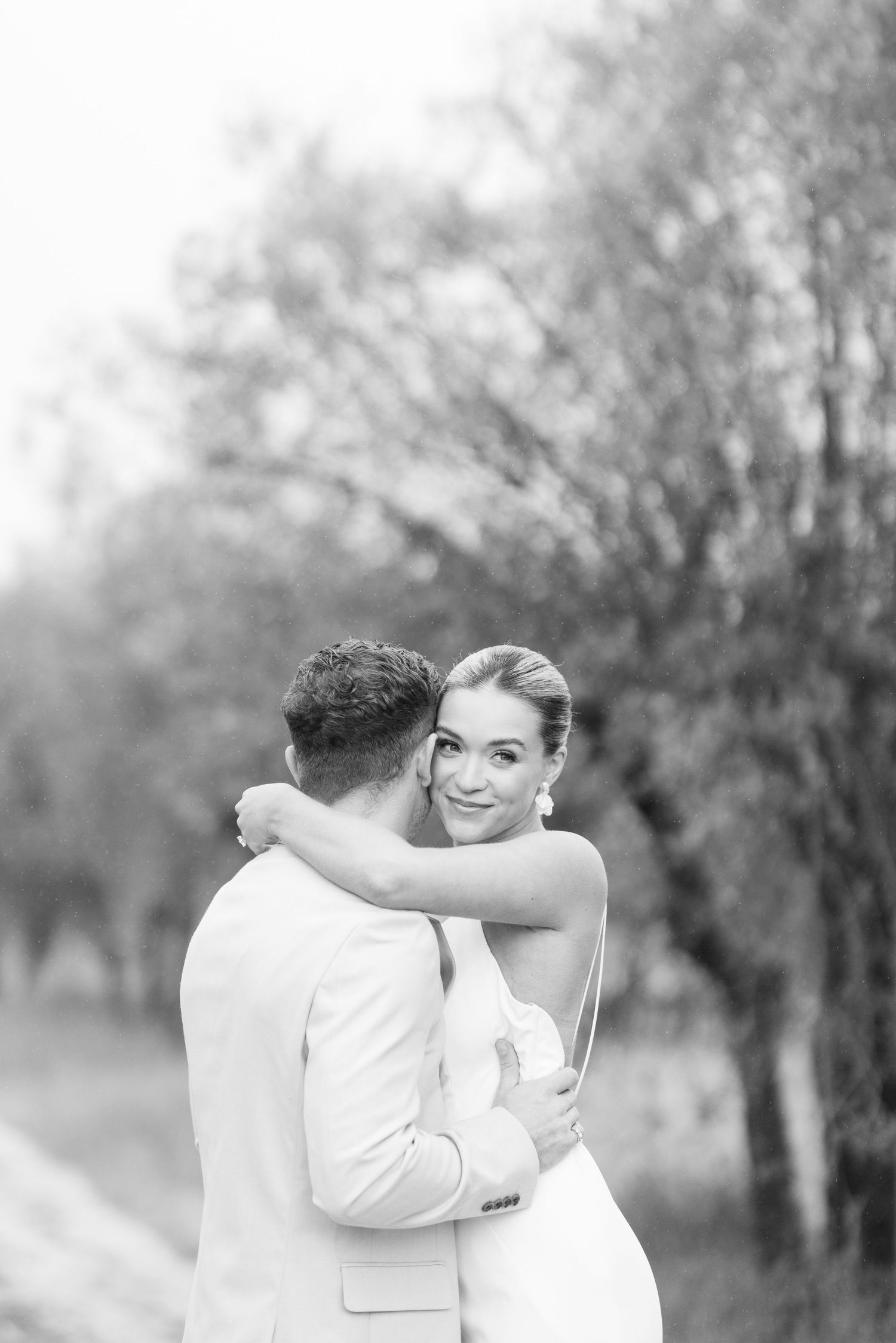Black and white photo of a couple embracing outdoors in a natural setting with trees in the background.