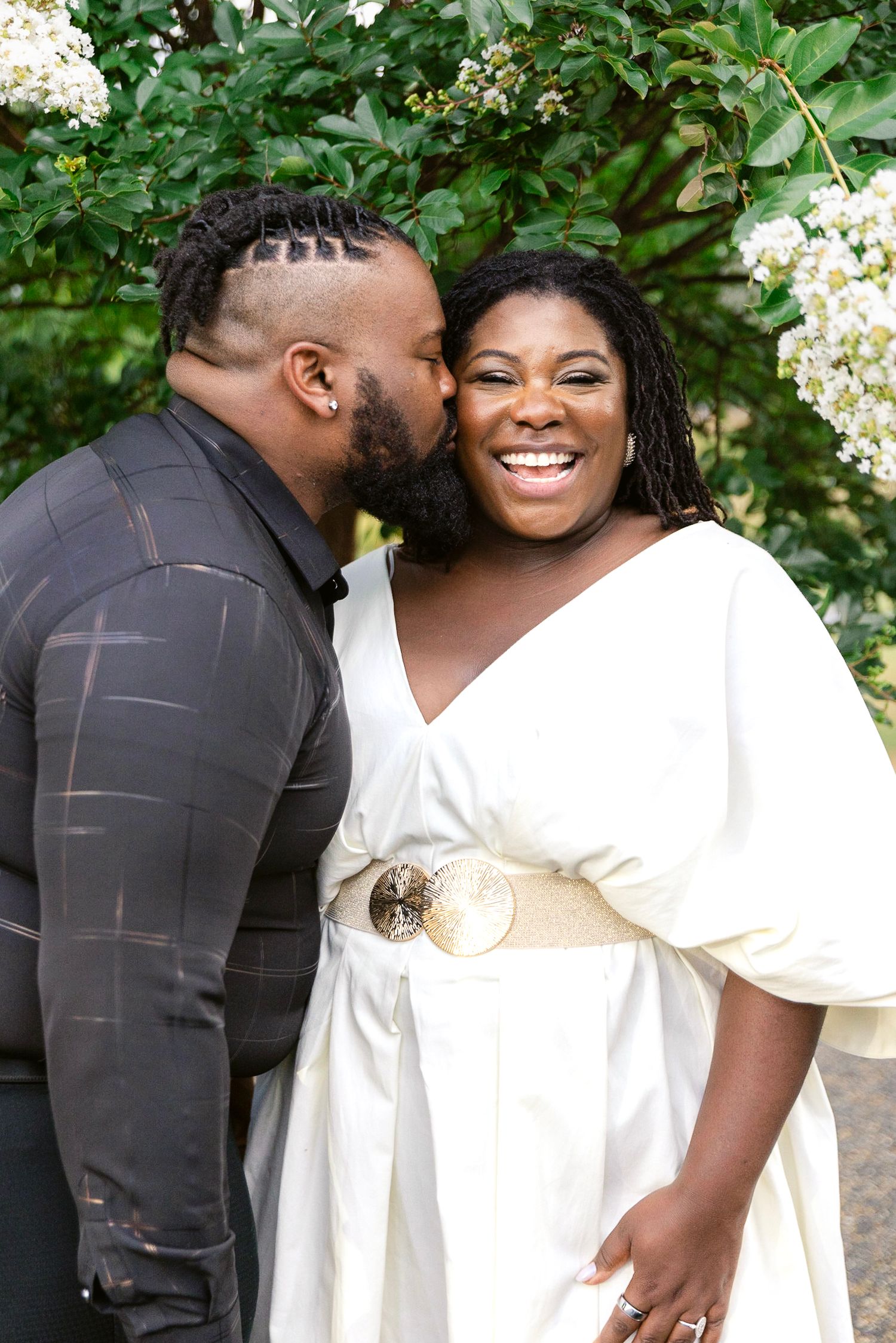 A romantic couple shares tender moments in a garden setting with white flowering bushes in the background.