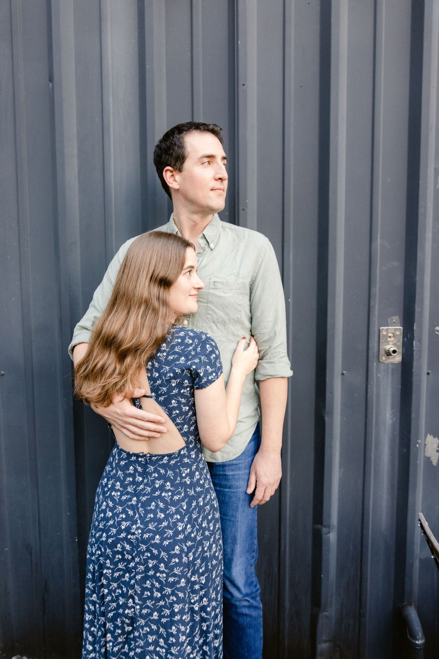 A couple embraces against a gray corrugated metal wall, with her wearing a blue floral dress and him in a light button-down shirt.