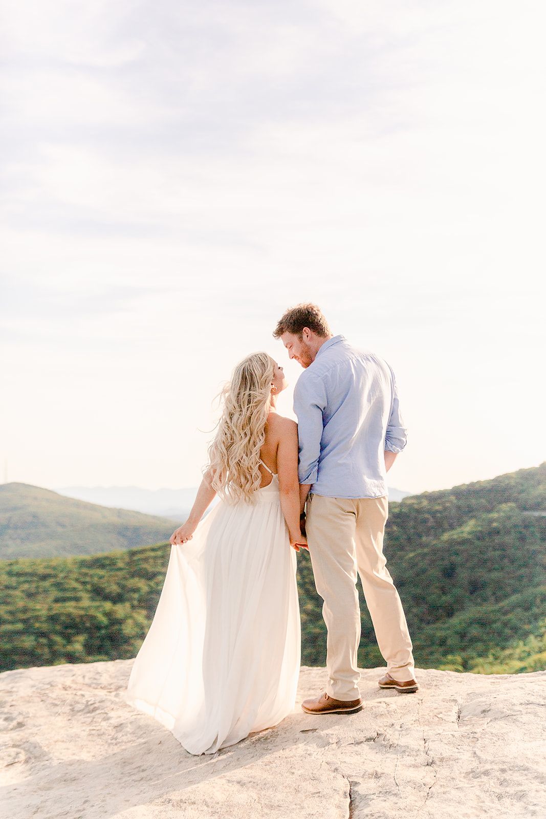 A couple in white wedding attire stands on a scenic mountain cliff overlooking lush green hills at sunset.