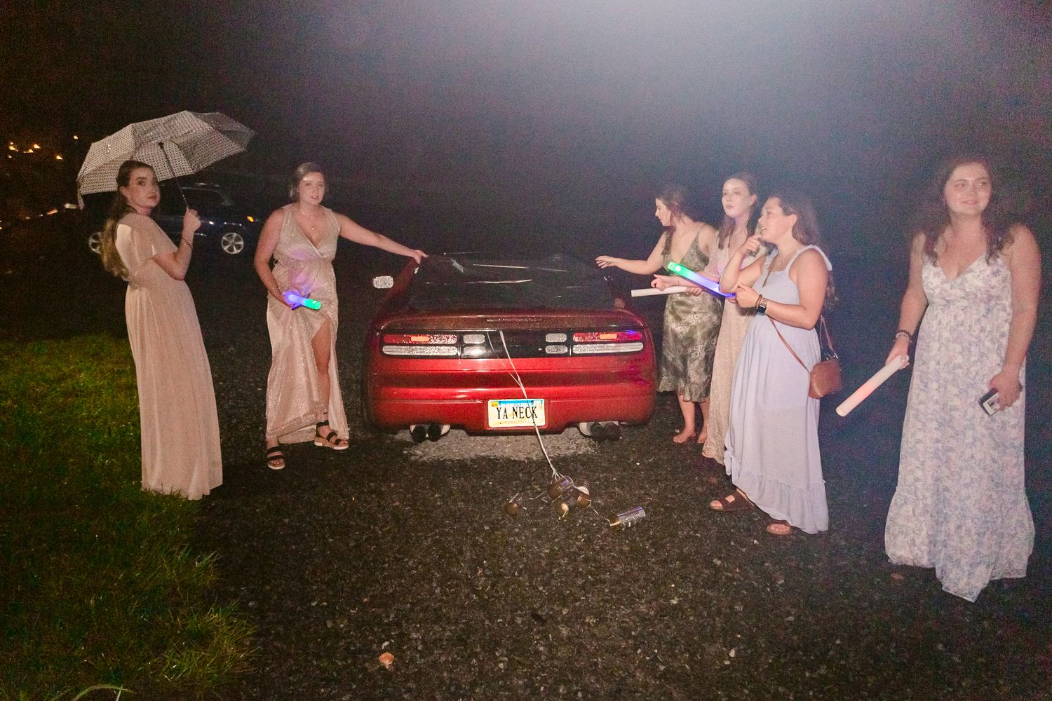 A group in light-colored dresses stands near a vintage red car at dusk during what appears to be a summer gathering.