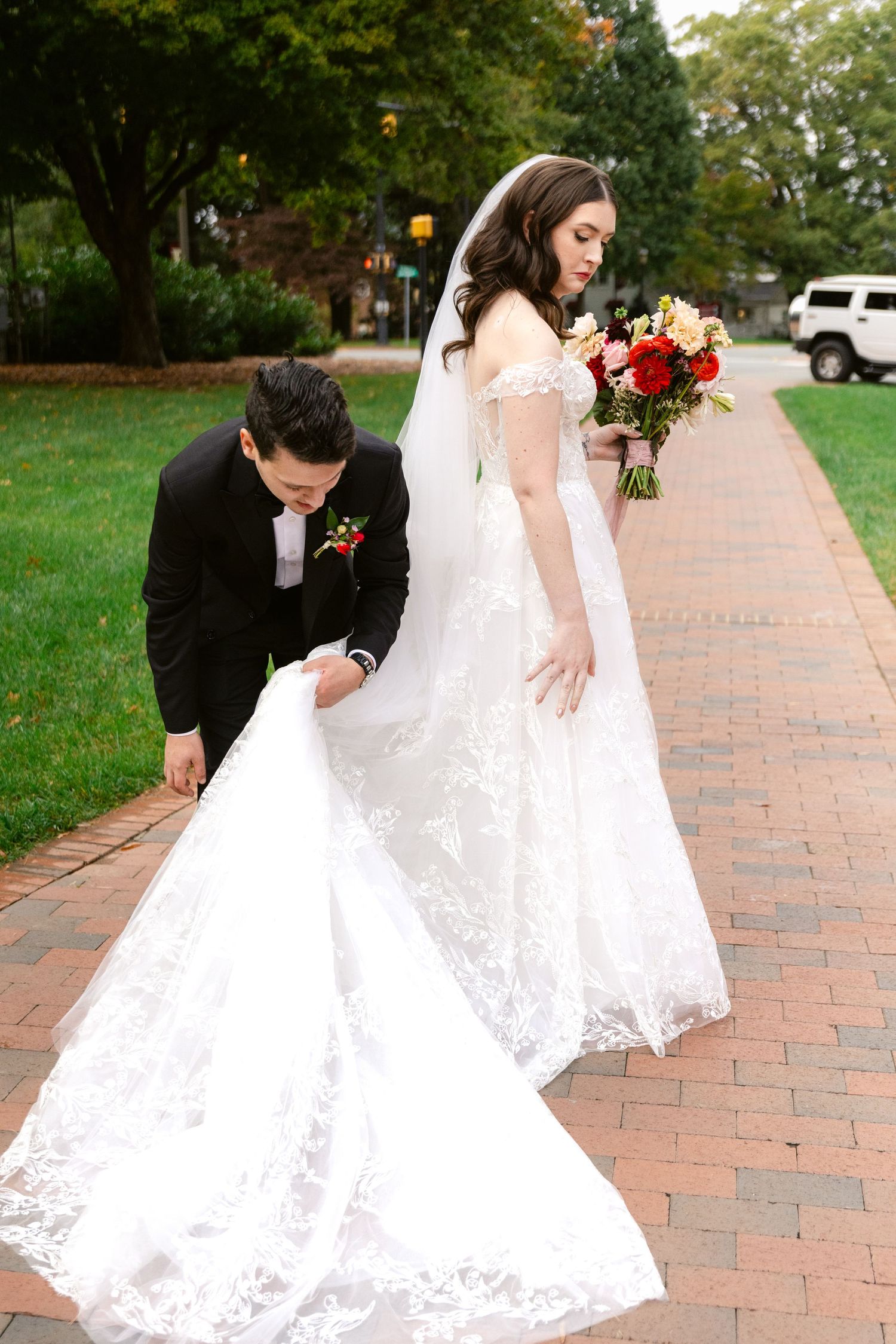 A couple in wedding attire on a brick path, with the groom adjusting the bride's long lace train.
