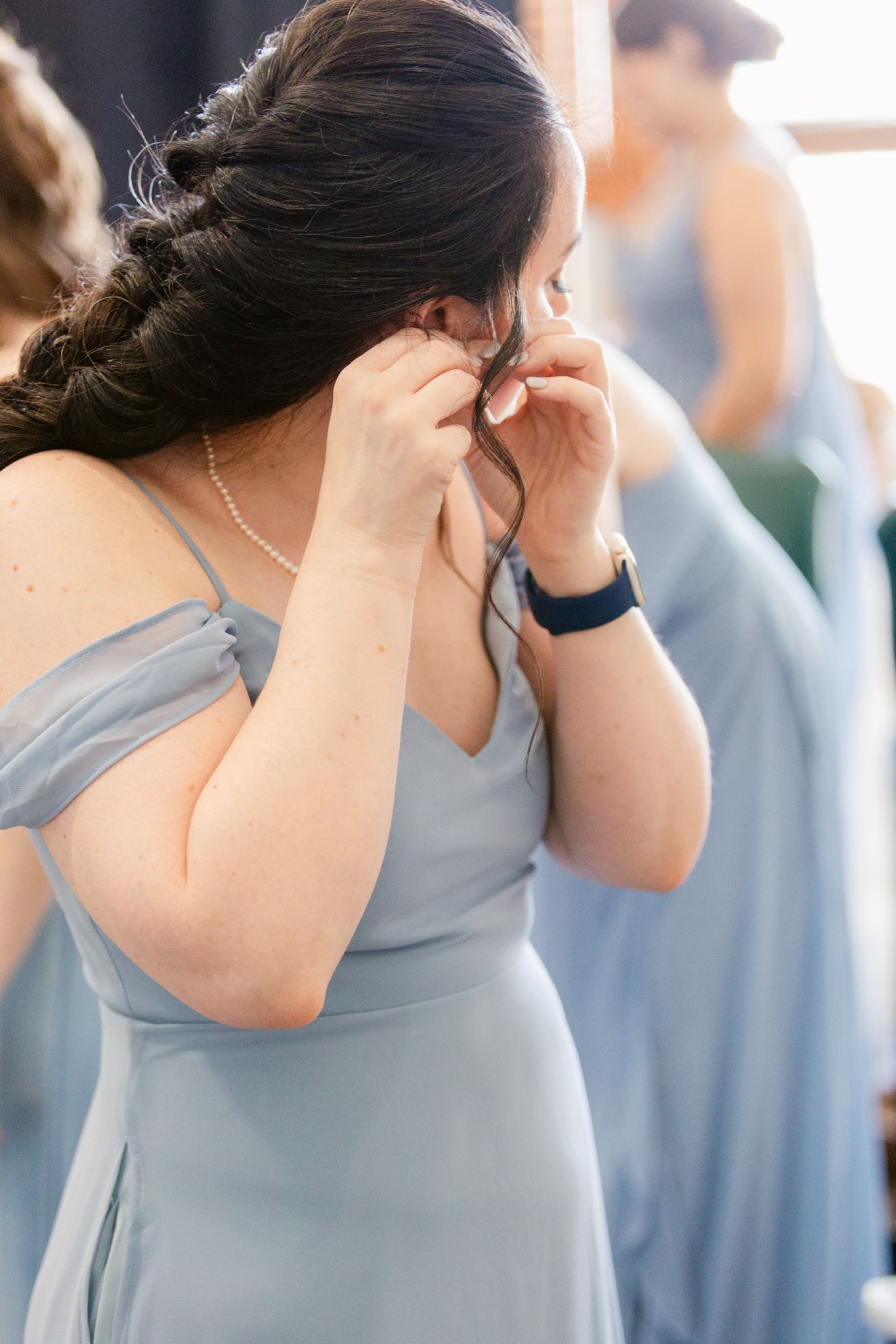 Woman in light blue bridesmaid dress puts on earrings during wedding preparations.