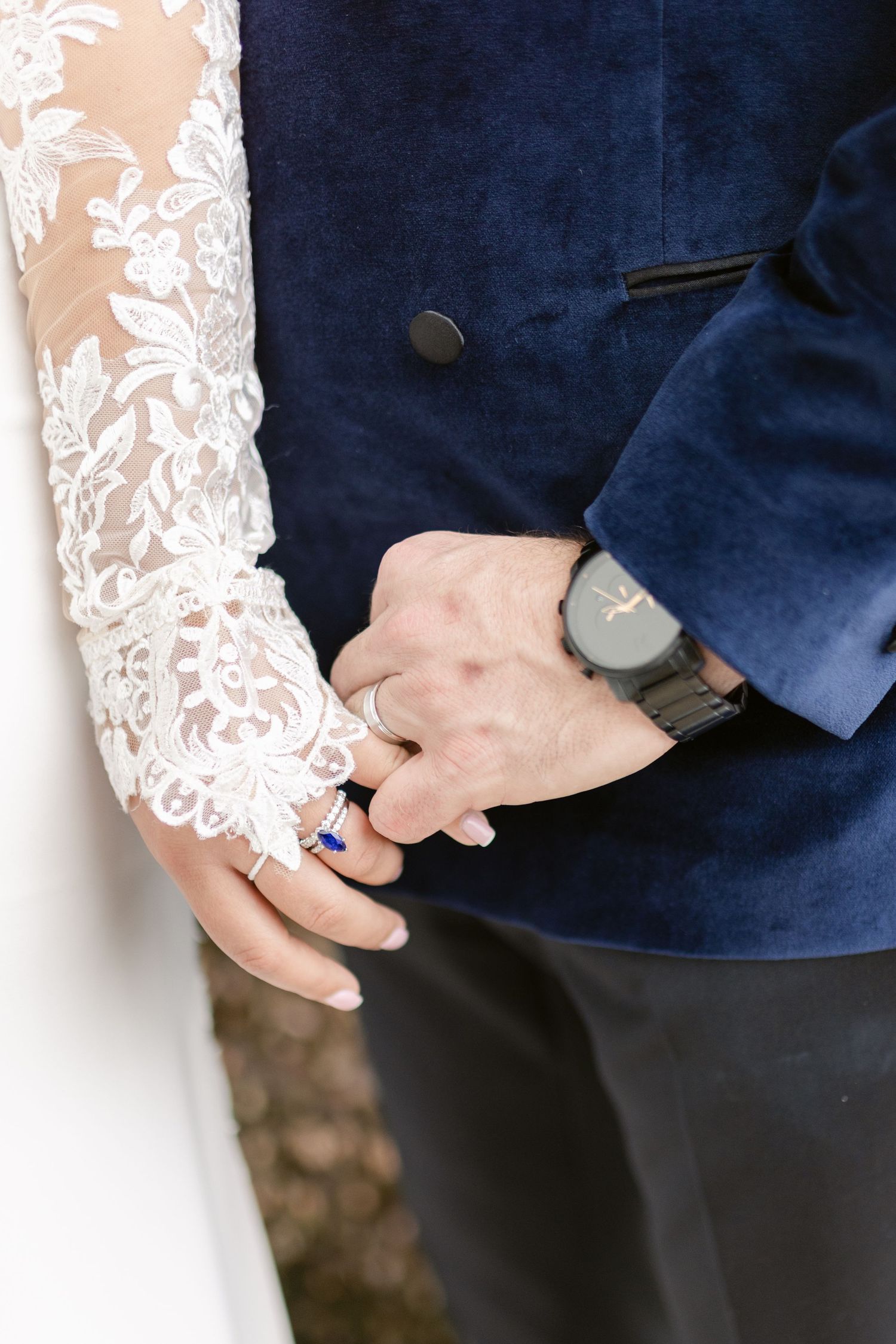 Close-up of two hands with wedding rings against a navy blue suit and lace dress sleeve.