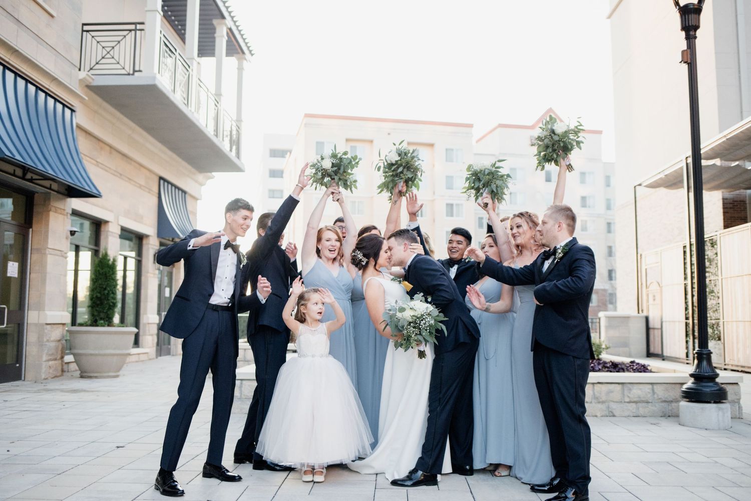 Wedding group celebrates with energetic poses and raised arms in outdoor setting.