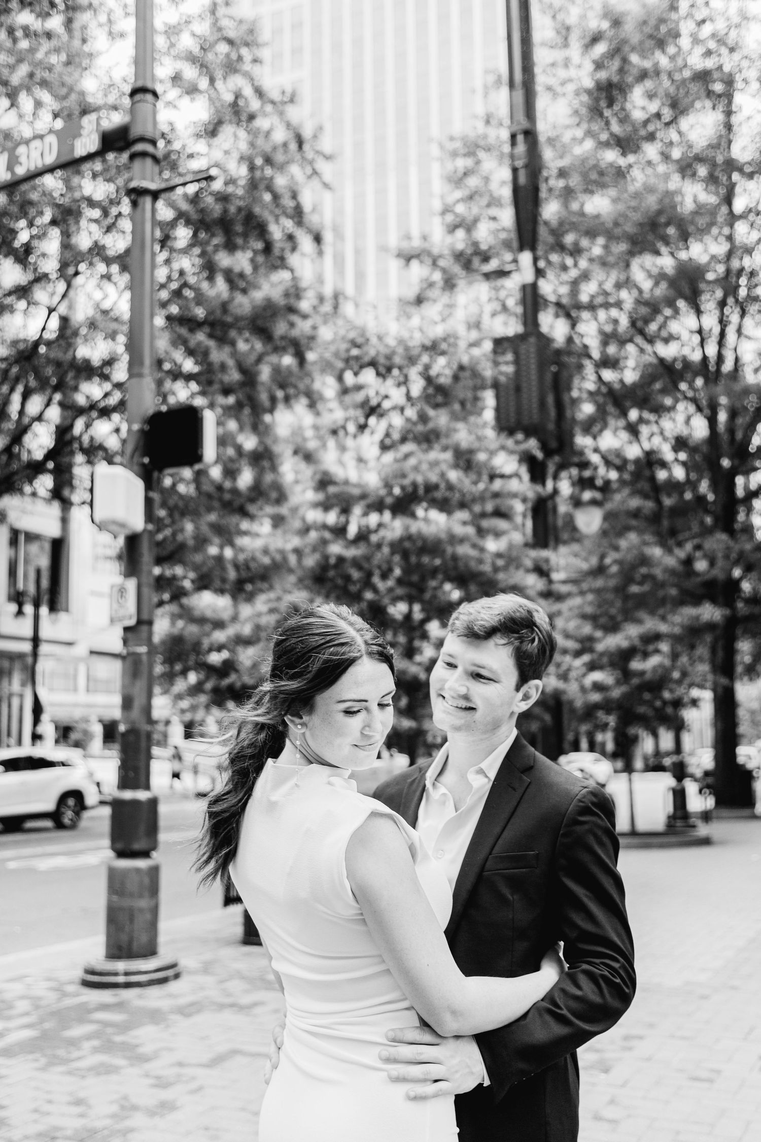 A romantic black and white portrait of a couple embracing on a city street with lamp posts and trees in the background.