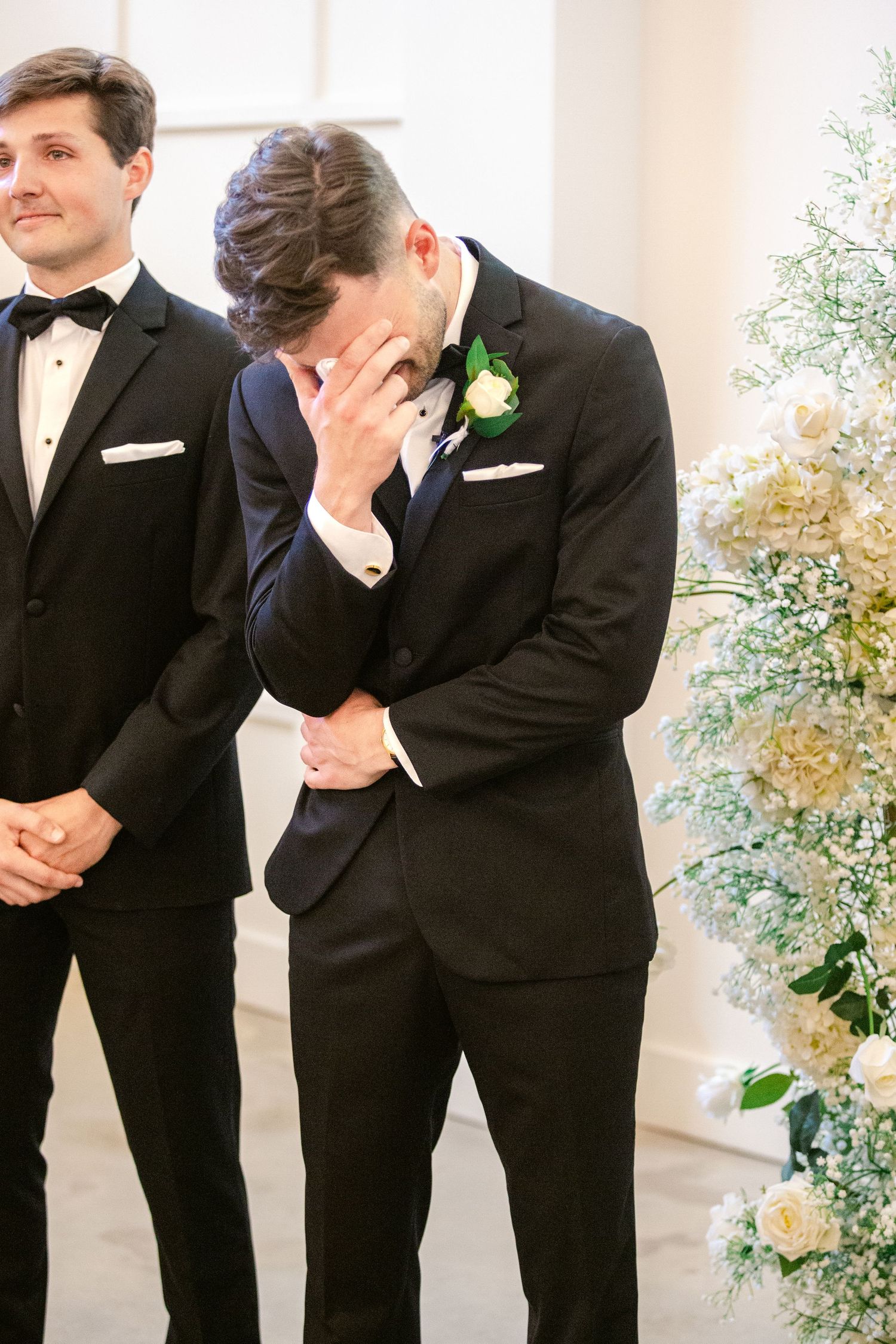 Two groomsmen in black tuxedos share an emotional moment at the altar during a wedding ceremony.