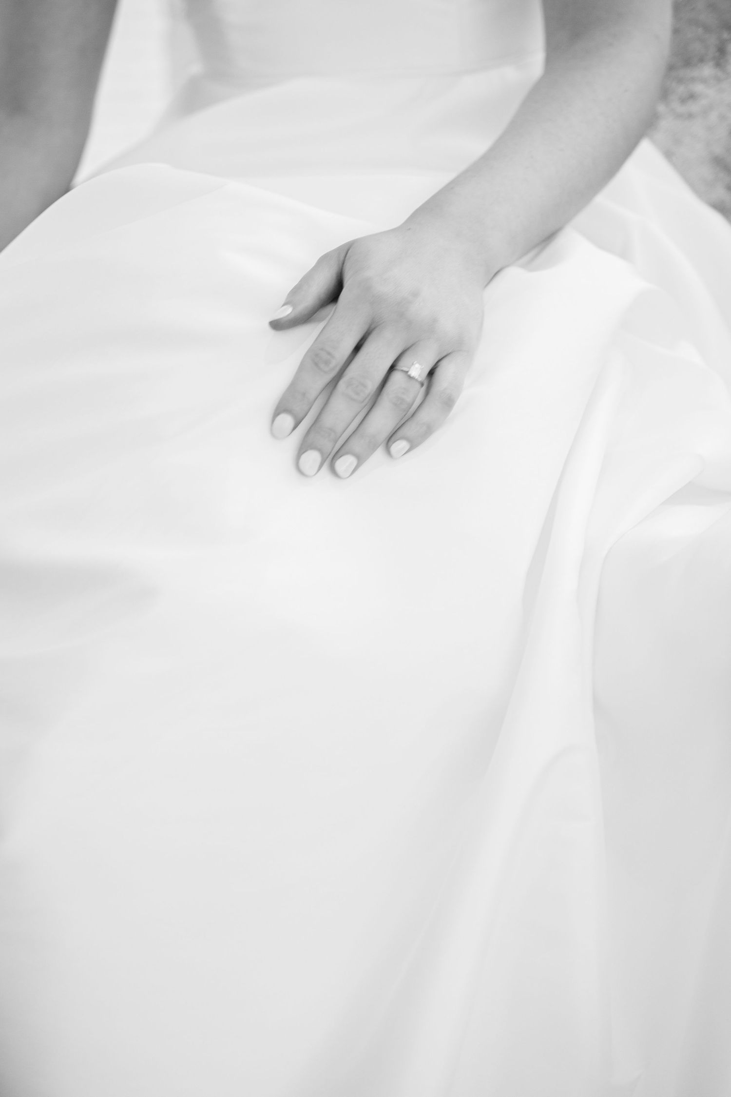 Close-up black and white photo of a hand with an engagement ring resting on white fabric.