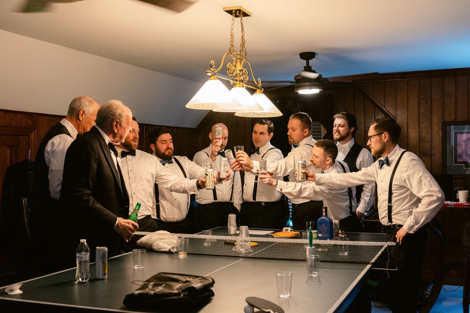 Groomsmen in formal attire gathered around ping pong table for pre-wedding celebration.