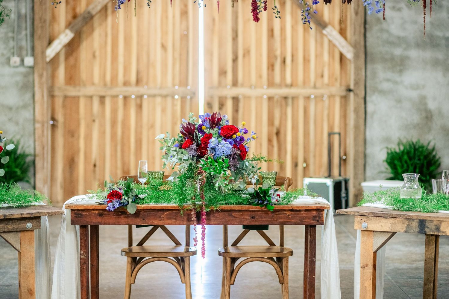 Sweetheart table setup with cross-back chairs and red floral arrangements against wooden backdrop.