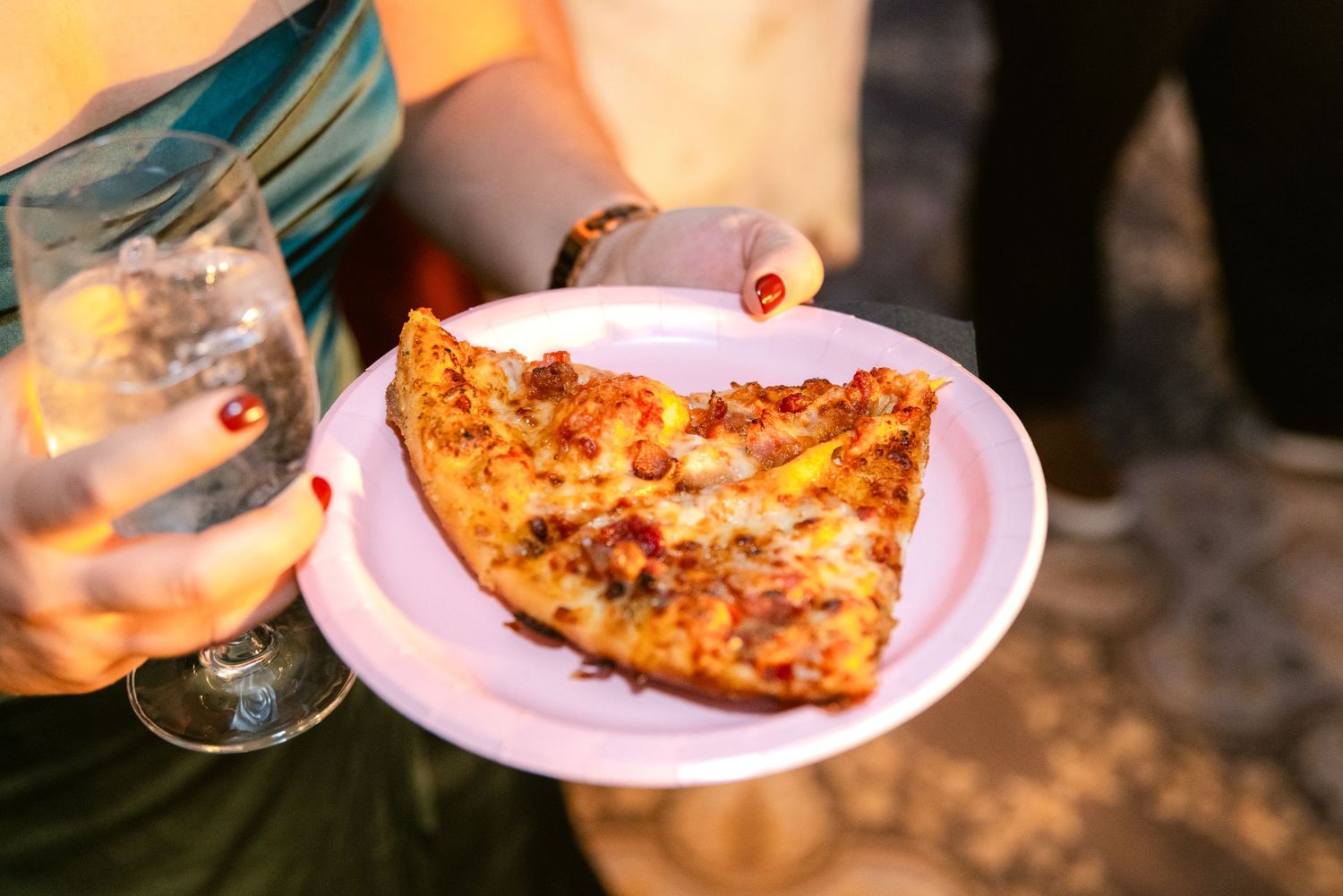 A slice of pepperoni pizza on a pink plate held against an orange sunset background with a glass visible.
