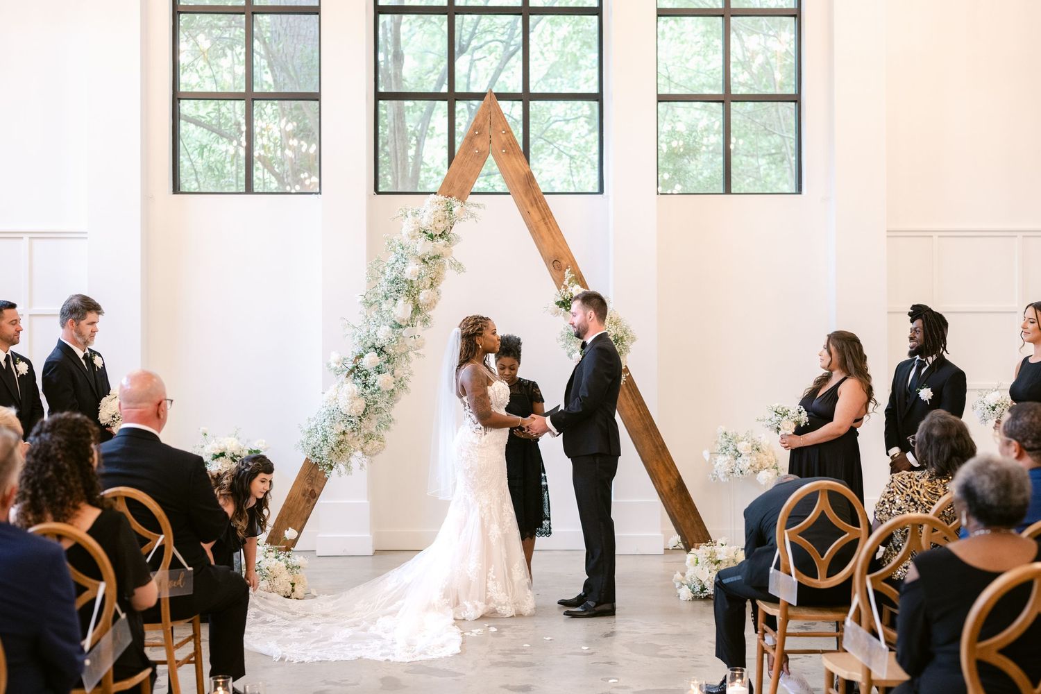 A wedding ceremony takes place in a bright white room with large windows and wooden cross-back chairs.