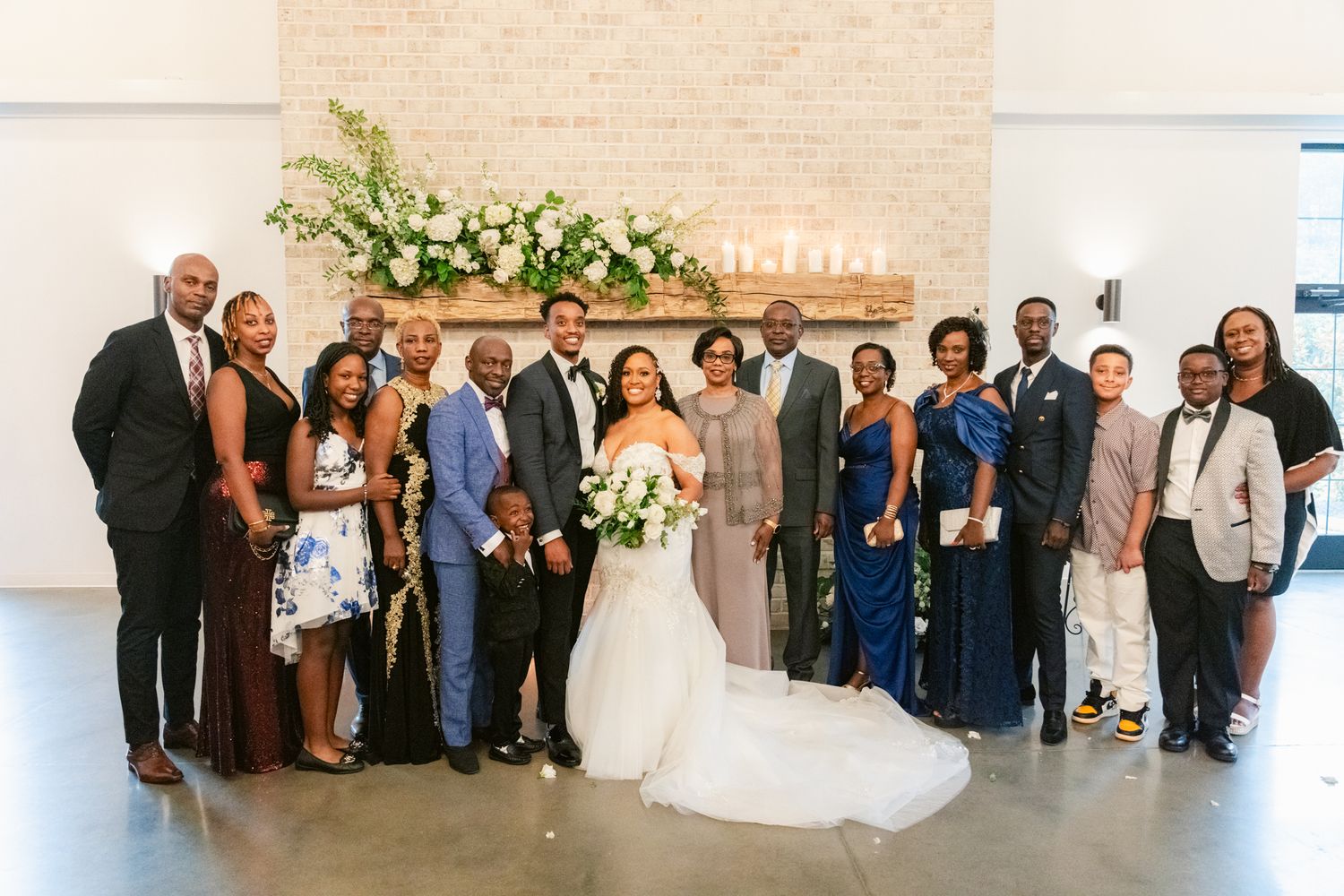 Wedding party group photo in front of white brick wall with floral arrangement at modern venue.