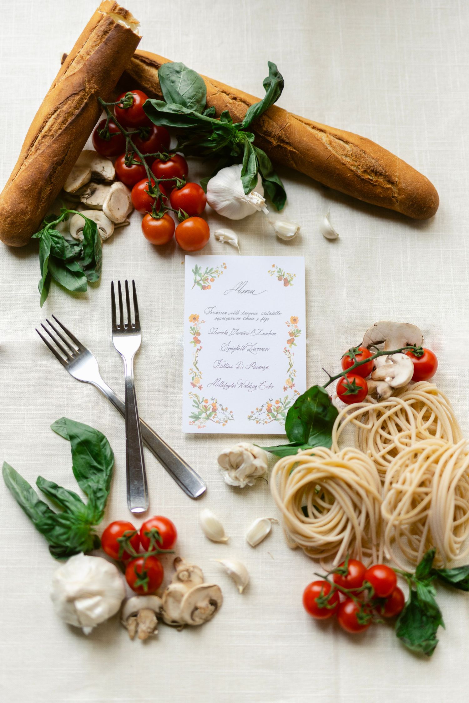 Fresh Italian cooking ingredients including pasta, cherry tomatoes, bread, basil and mushrooms arranged around an elegant menu card.