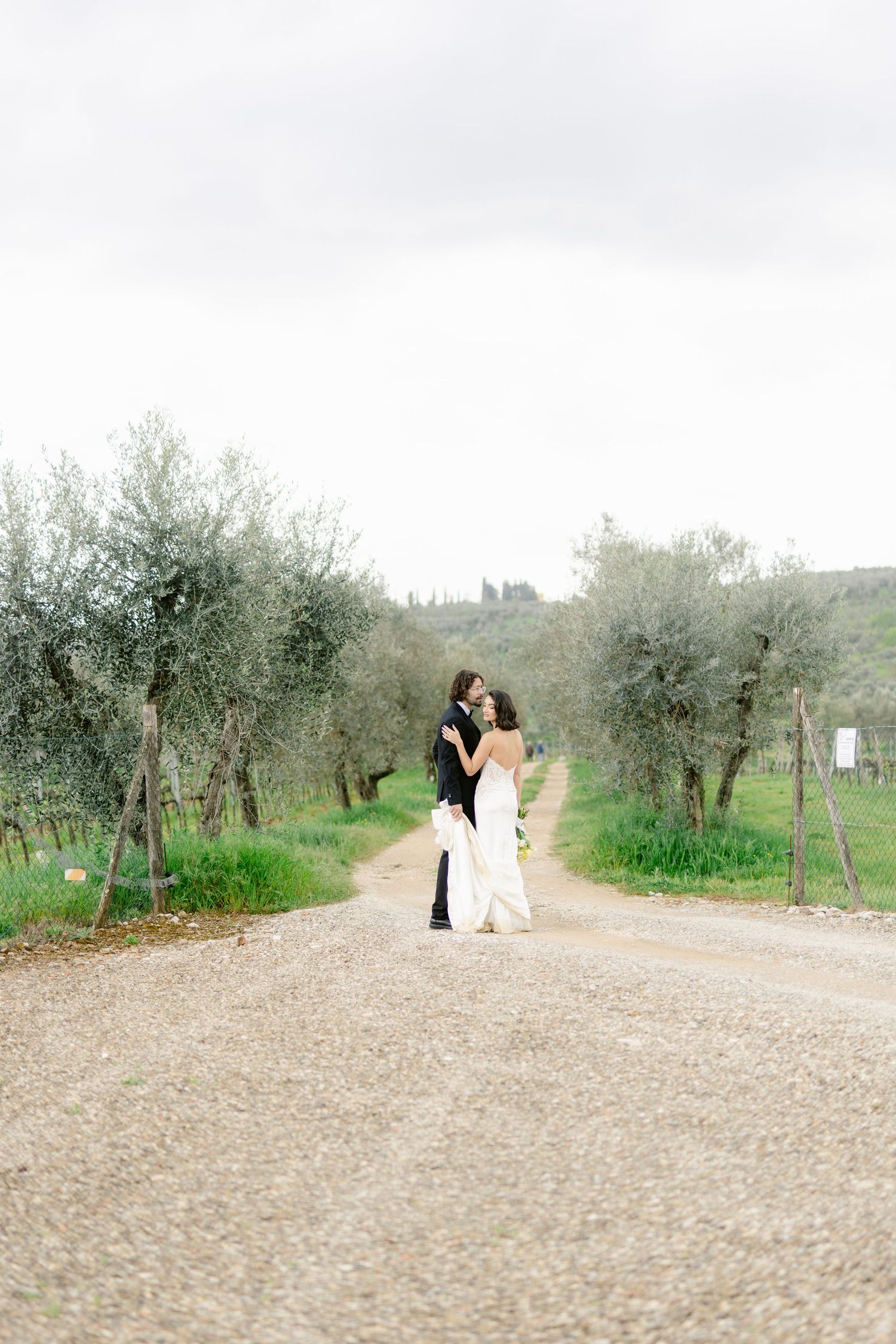 A romantic couple shares an intimate moment on a gravel path surrounded by olive trees in the countryside.