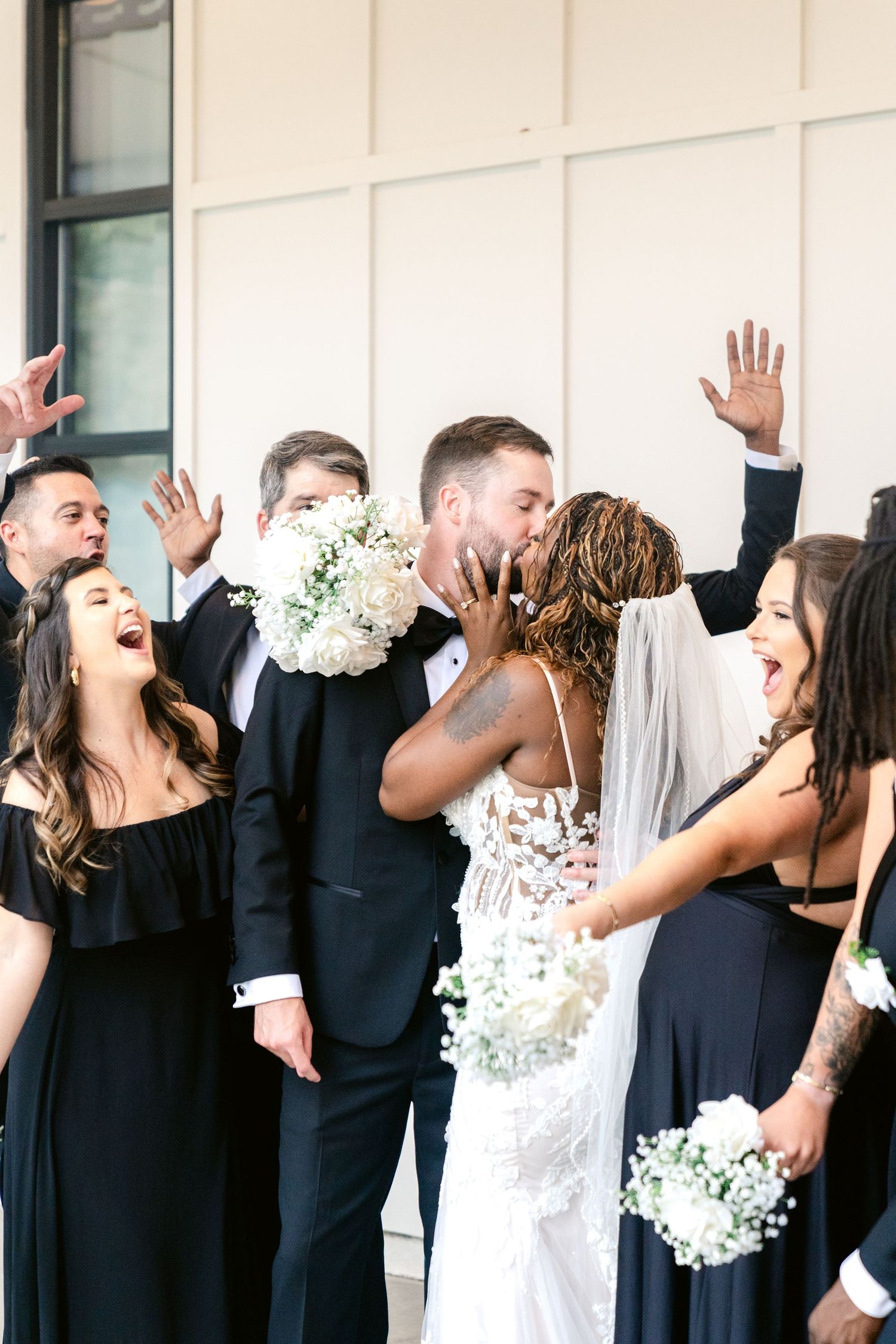 Wedding party in black and white attire poses for a fun jumping photo against a light colored wall.