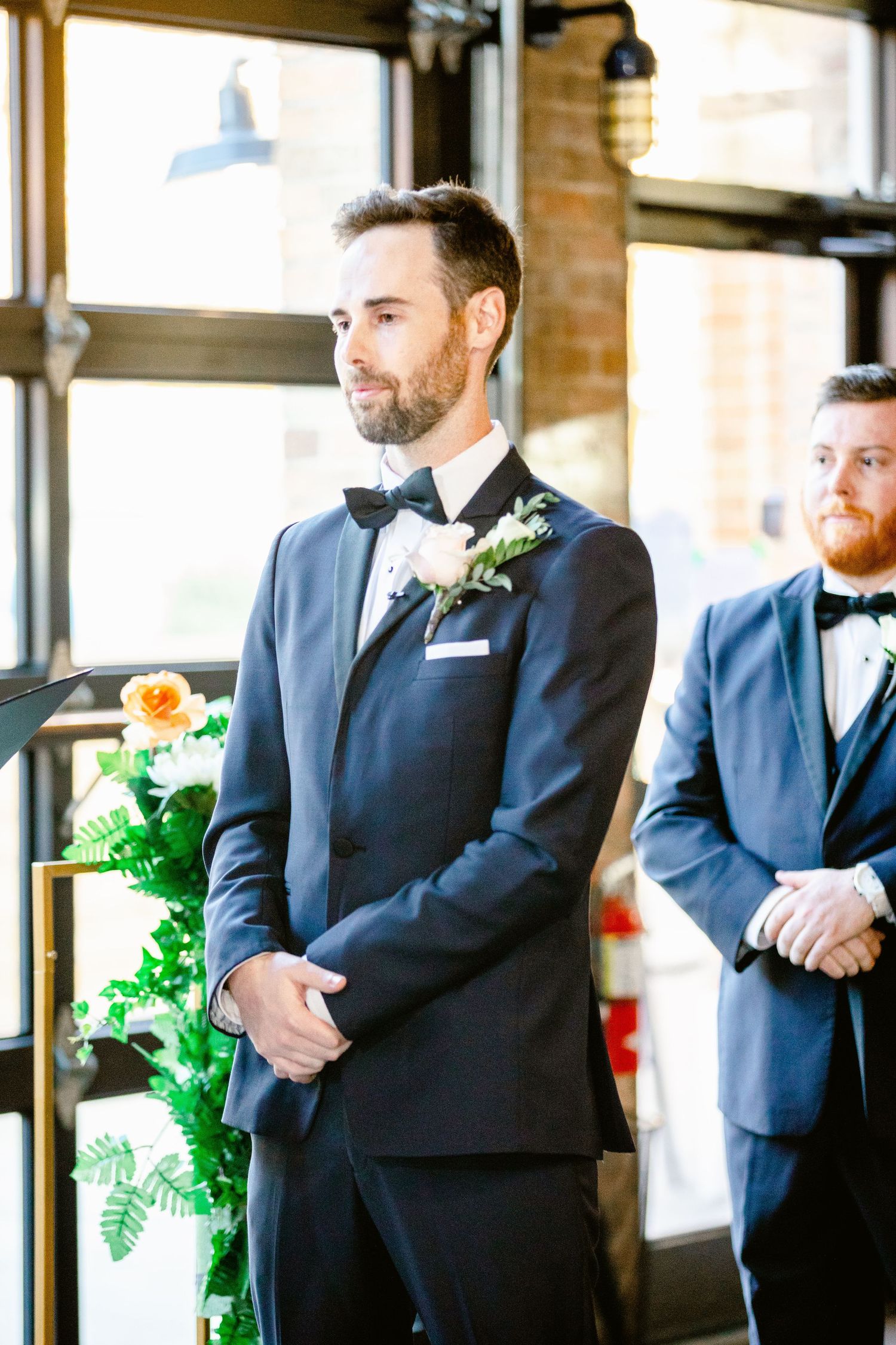 Groomsmen in matching navy blue suits stand in a row at an indoor wedding venue with natural lighting.