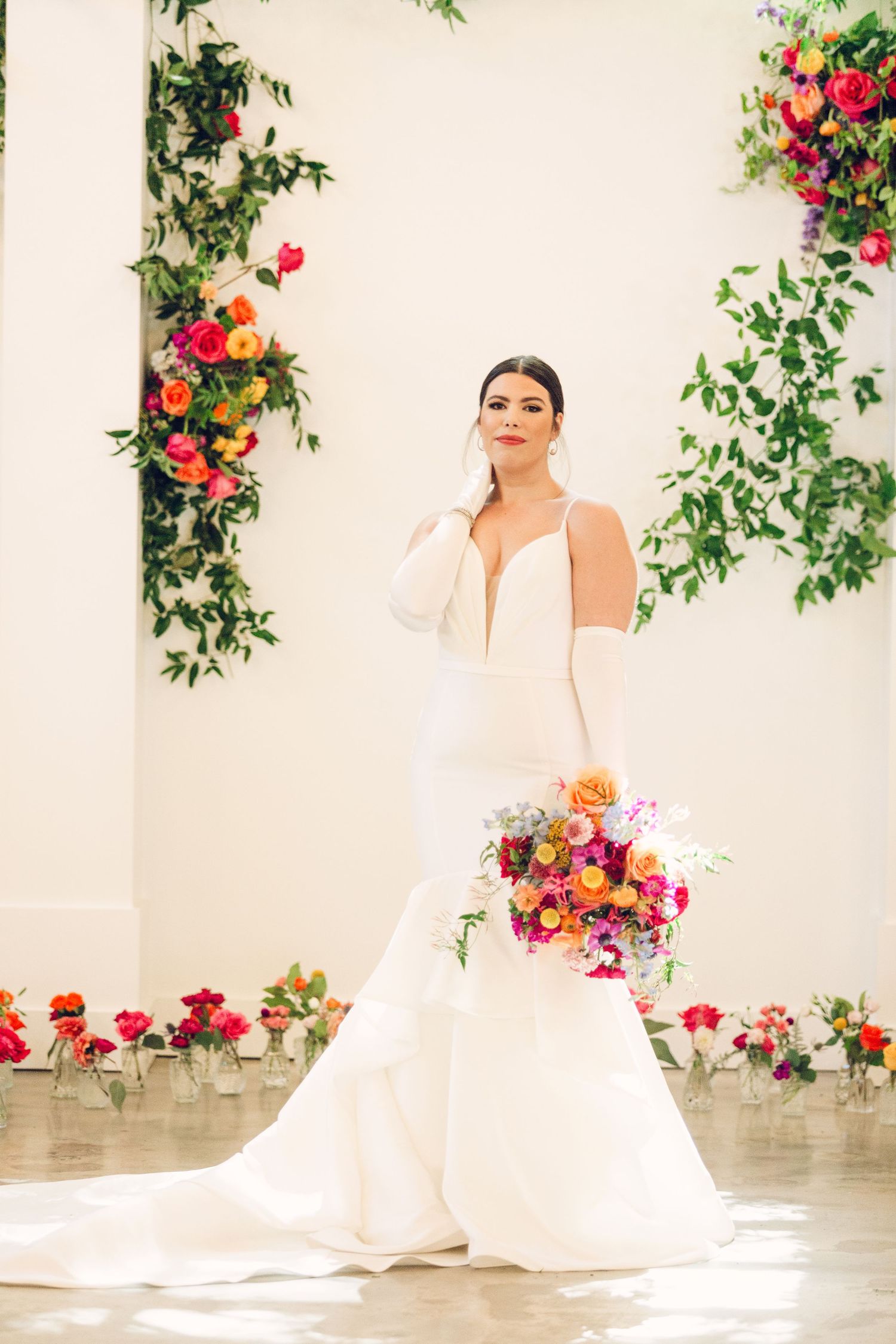 Bride in elegant white wedding dress poses gracefully against floral backdrop with pink and red roses.