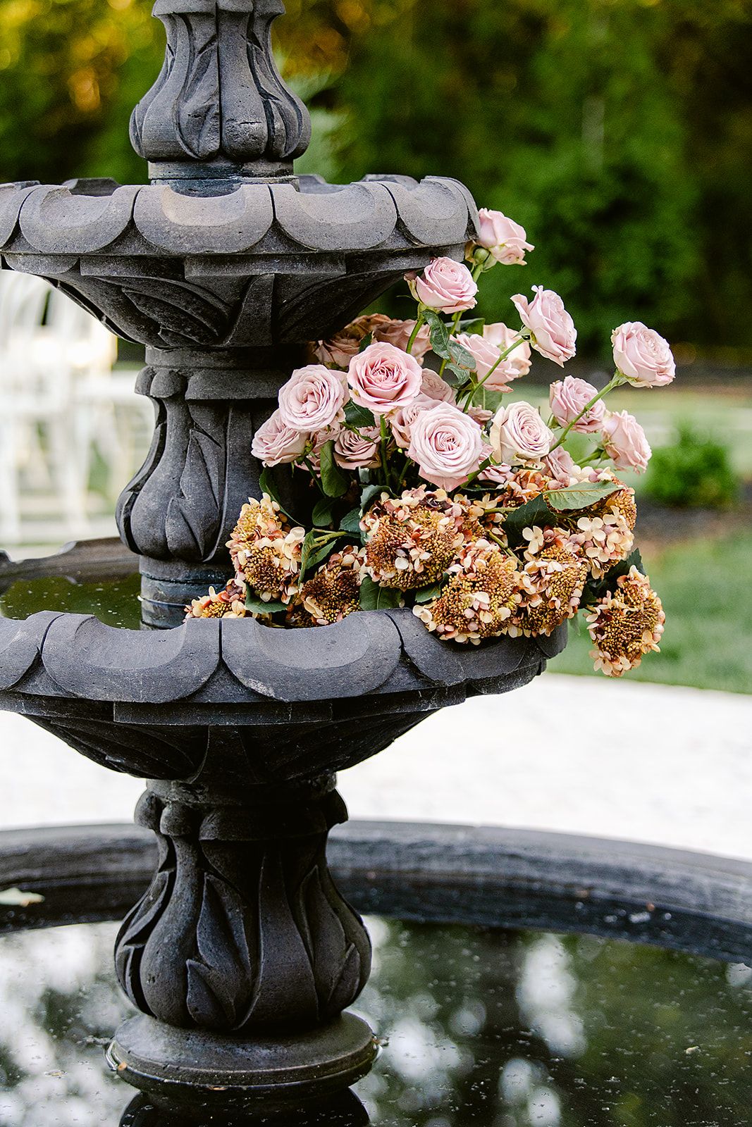 Pink roses and dried hydrangeas adorn a dark stone fountain in an outdoor garden setting.
