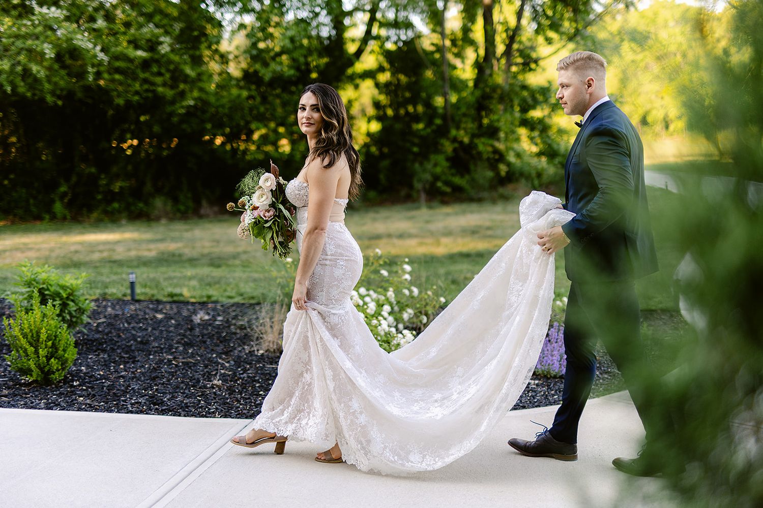 A couple in wedding attire walks together through a lush green garden setting during a summer outdoor ceremony.
