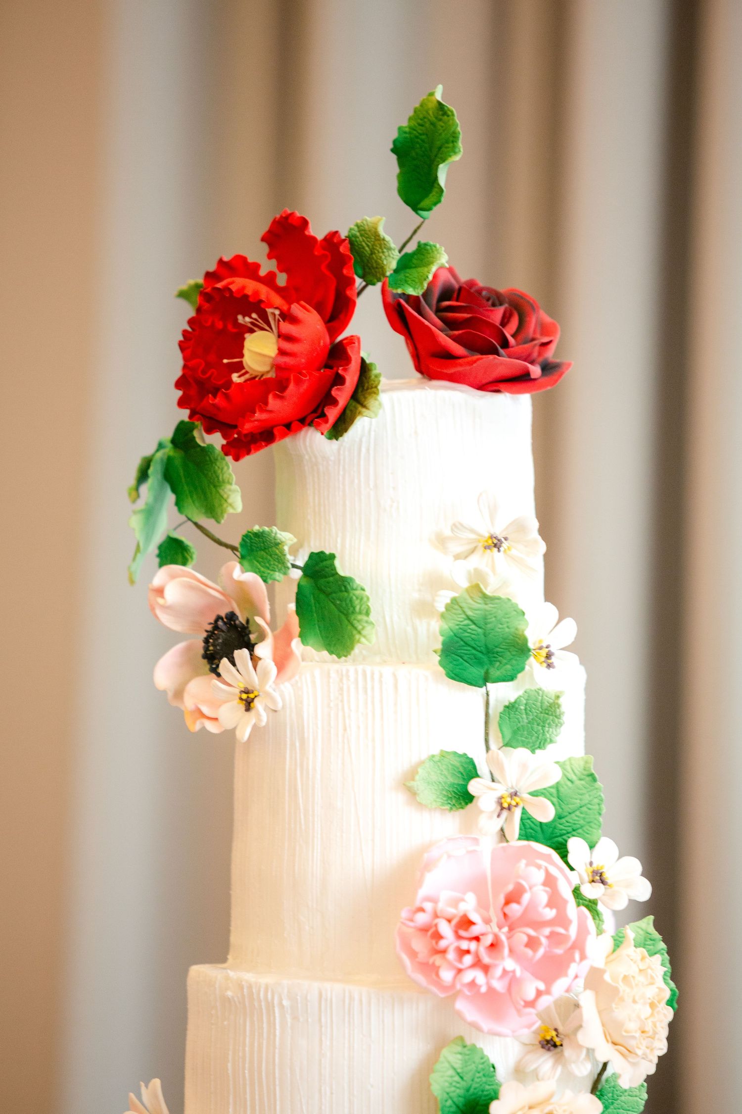 White wedding cake decorated with cascading red, pink and peach sugar flowers and green leaves on a neutral background.