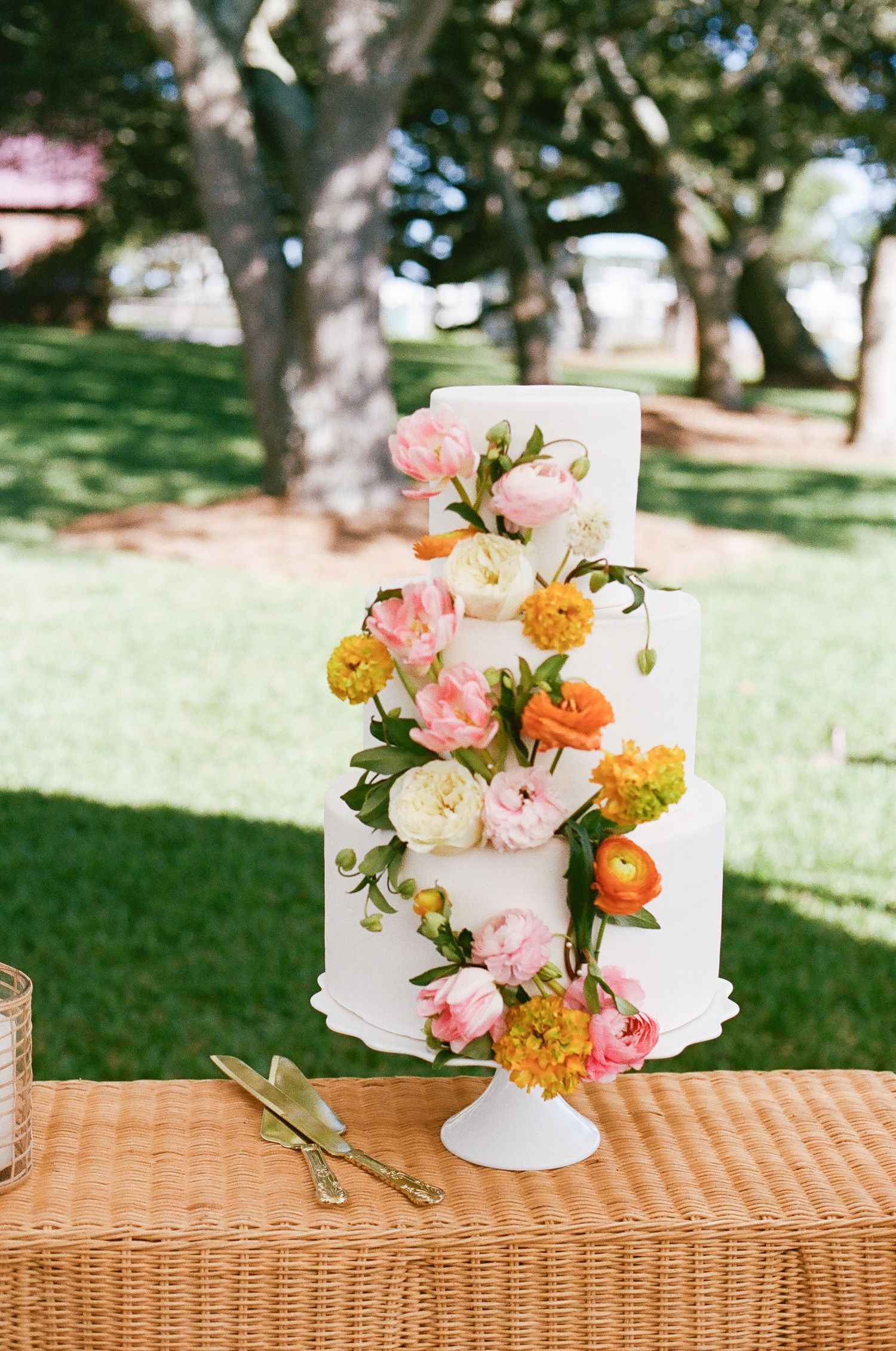 A three-tiered white wedding cake decorated with pink, orange and yellow garden roses on a wicker table.