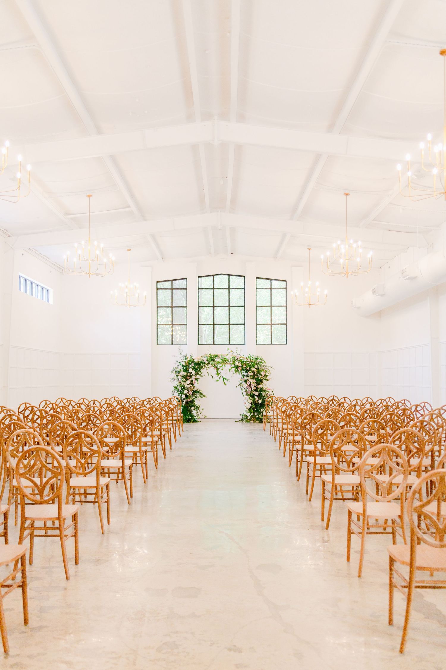 Rows of wooden chairs arranged in a bright white venue with windows and floral decorations for a wedding ceremony.