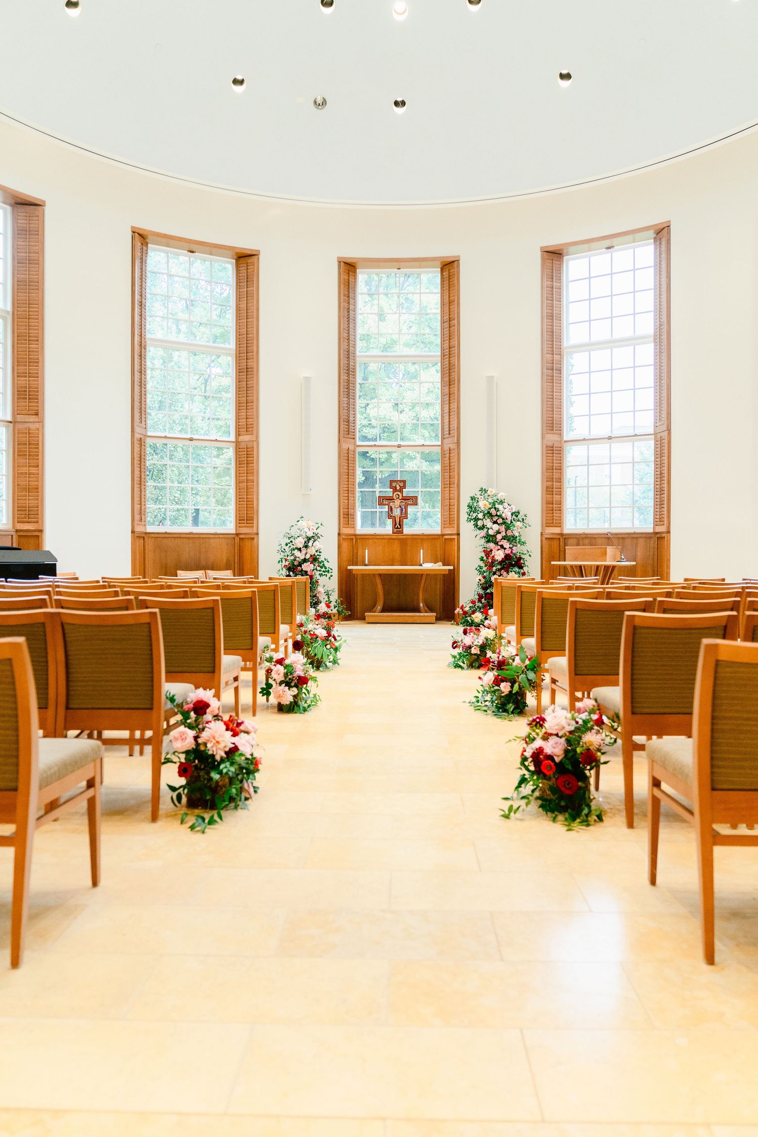 An elegant chapel interior with tall windows and floral arrangements lining the aisle for a wedding ceremony.