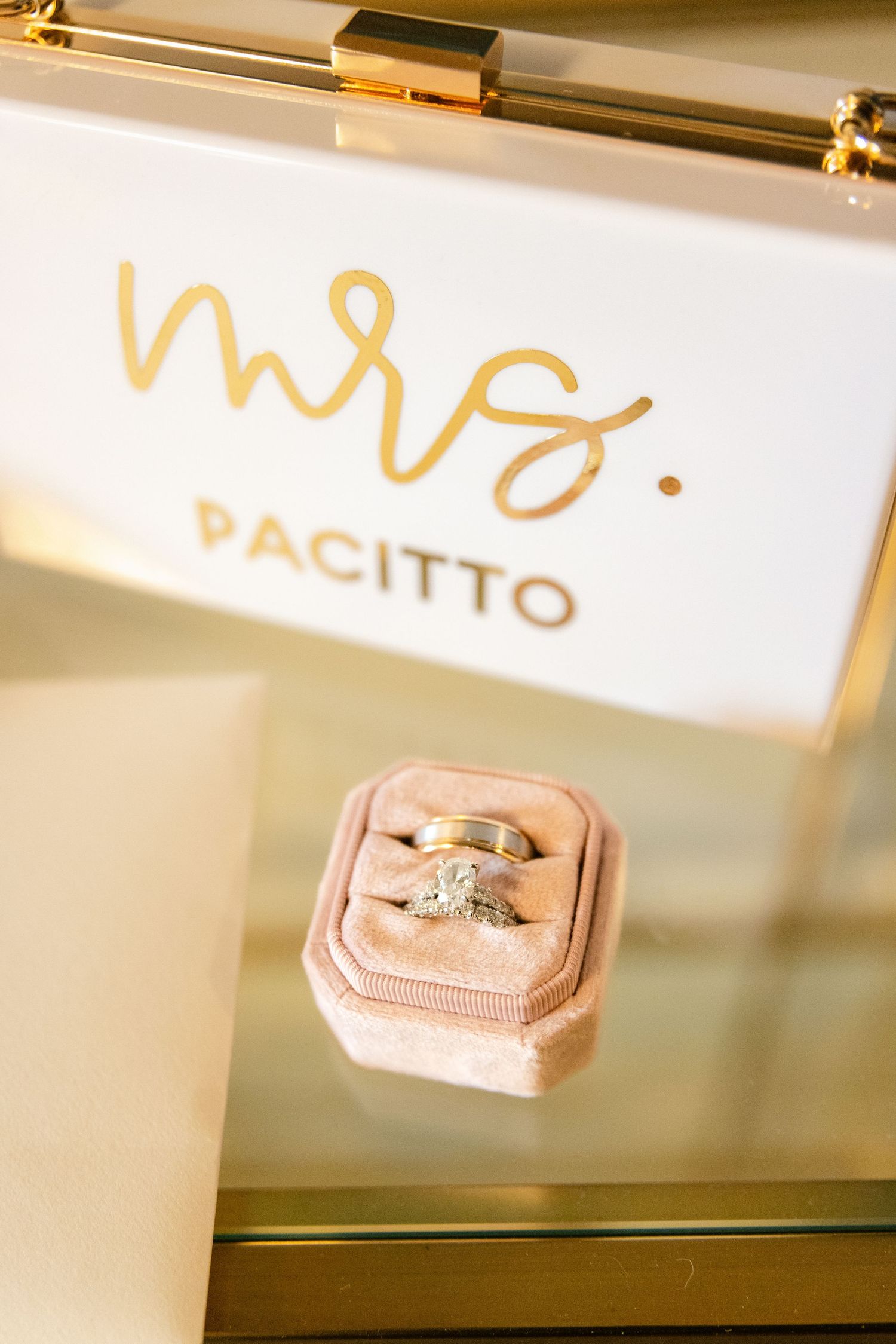 Gold wedding calligraphy text 'Mrs. Pacitto' displayed above a pink velvet ring box on a reflective surface.
