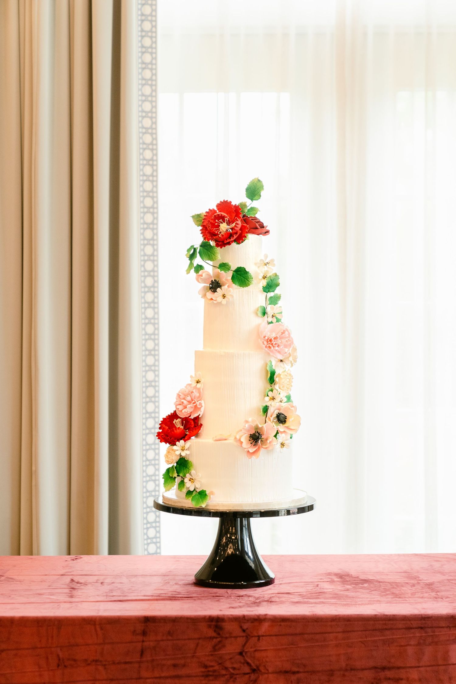Elegant white wedding cake with coral and red floral decorations displayed on a black pedestal against a light background.